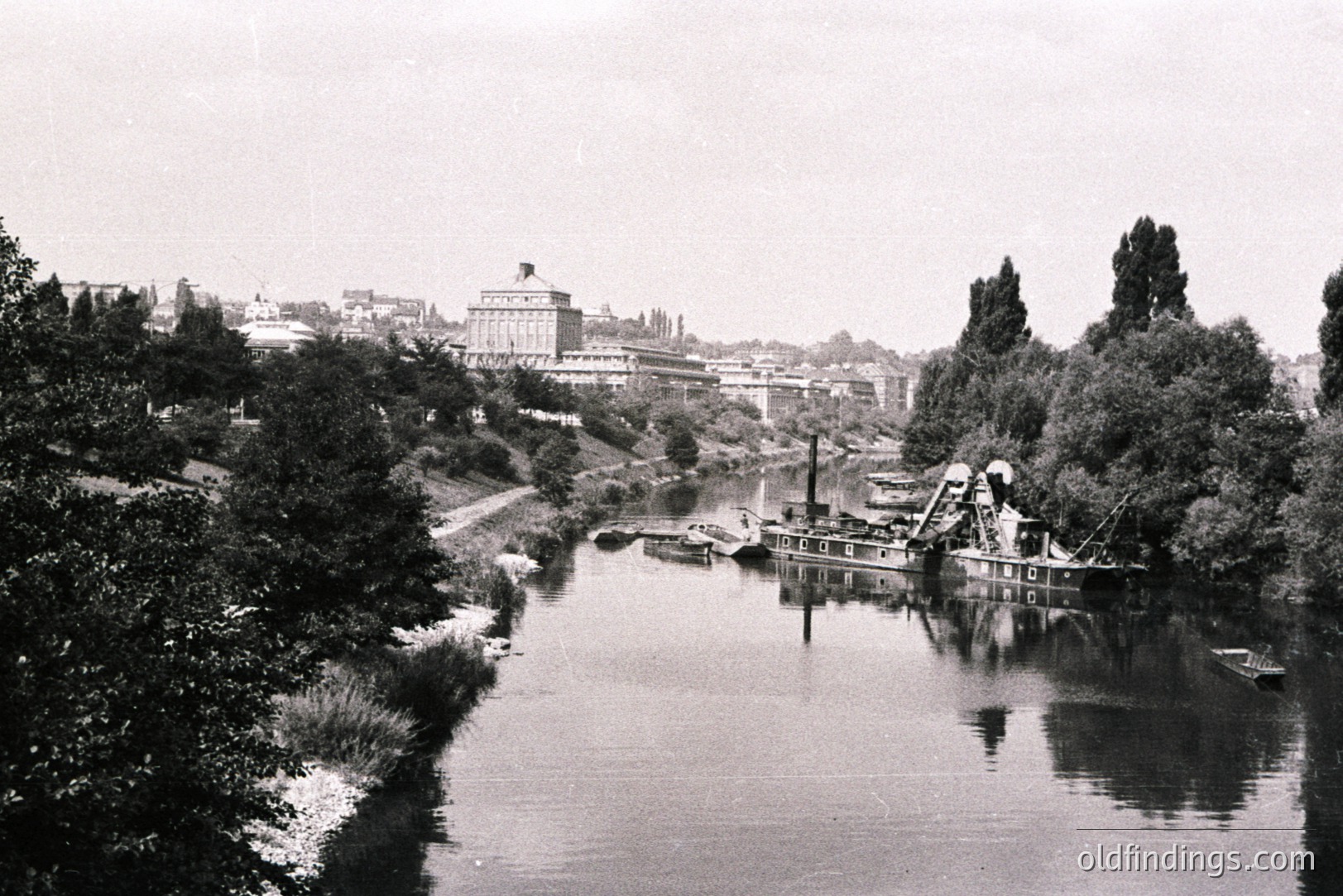 A wide river reflects a large, ornate building with a central tower and surrounding smaller structures. A dredging barge occupies the waterway, suggesting industrial activity. Lush vegetation lines the riverbanks, framing the scene. Likely a European river city, early 20th century.