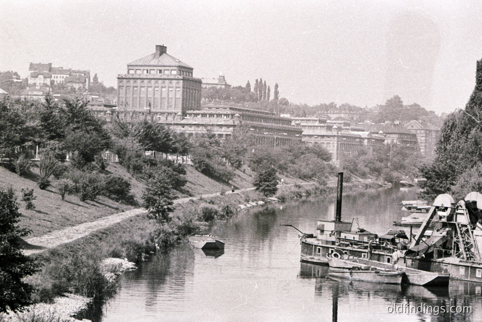 A long view of a river, likely the Rhine, with a large industrial building complex dominating the hillside. A dredging boat is moored in the foreground. Likely late 19th or early 20th century, based on clothing & technology. Potential industrial heritage site.