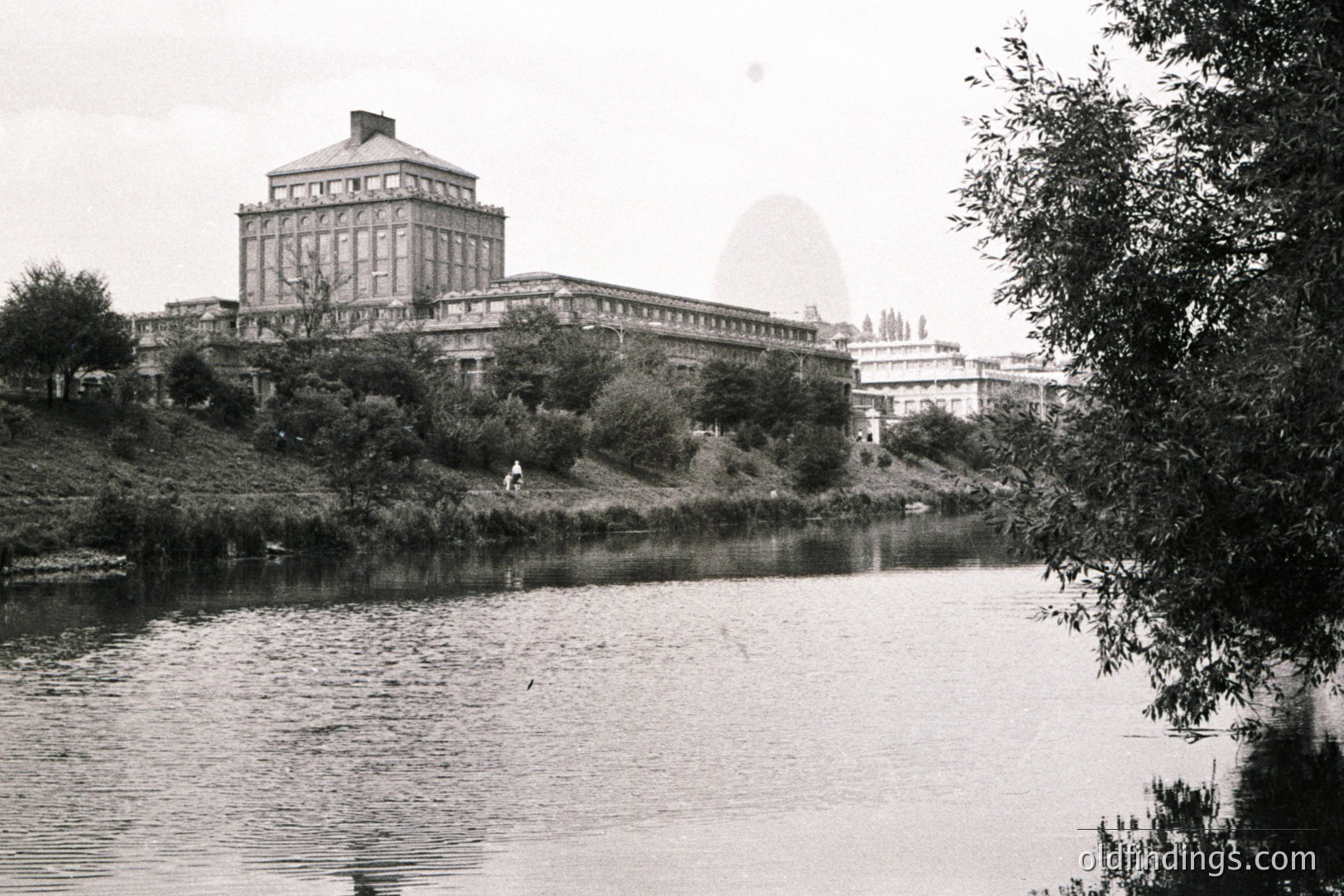 A stately, neoclassical building overlooks a still river, likely a resort or sanatorium. The building features a central cupola & symmetrical facade. A figure walks along a distant path. Photograph exhibits characteristics of mid-century documentation, possibly 1950s-1970s. Appears to be in a European setting.