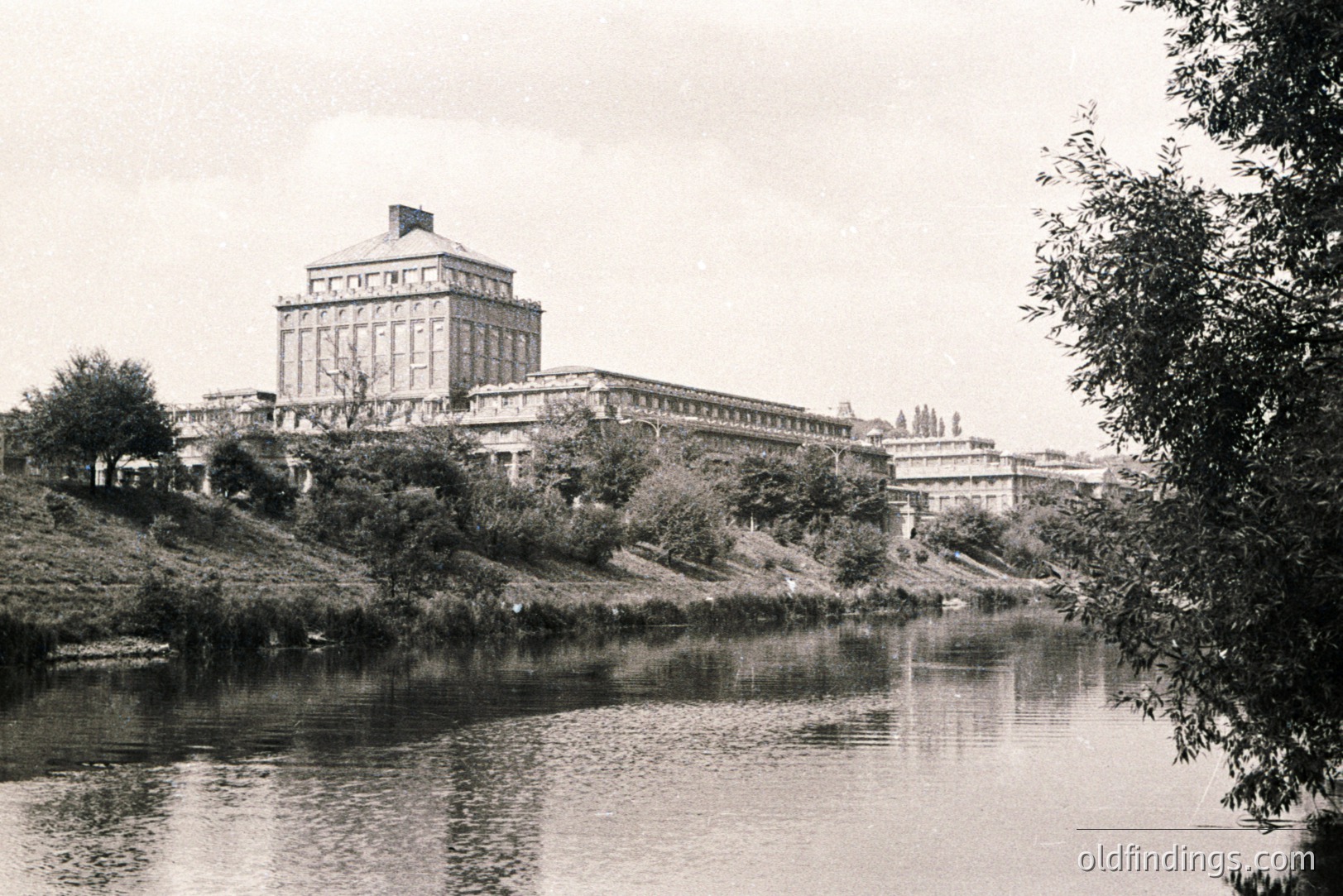 Grand, imposing institutional building with classical details rises above a riverbank. Multi-story structure features a prominent central tower, likely a library or administrative center. Likely a university or government complex. Architectural style suggests early 20th century. Landscape includes grassy slopes & mature trees.