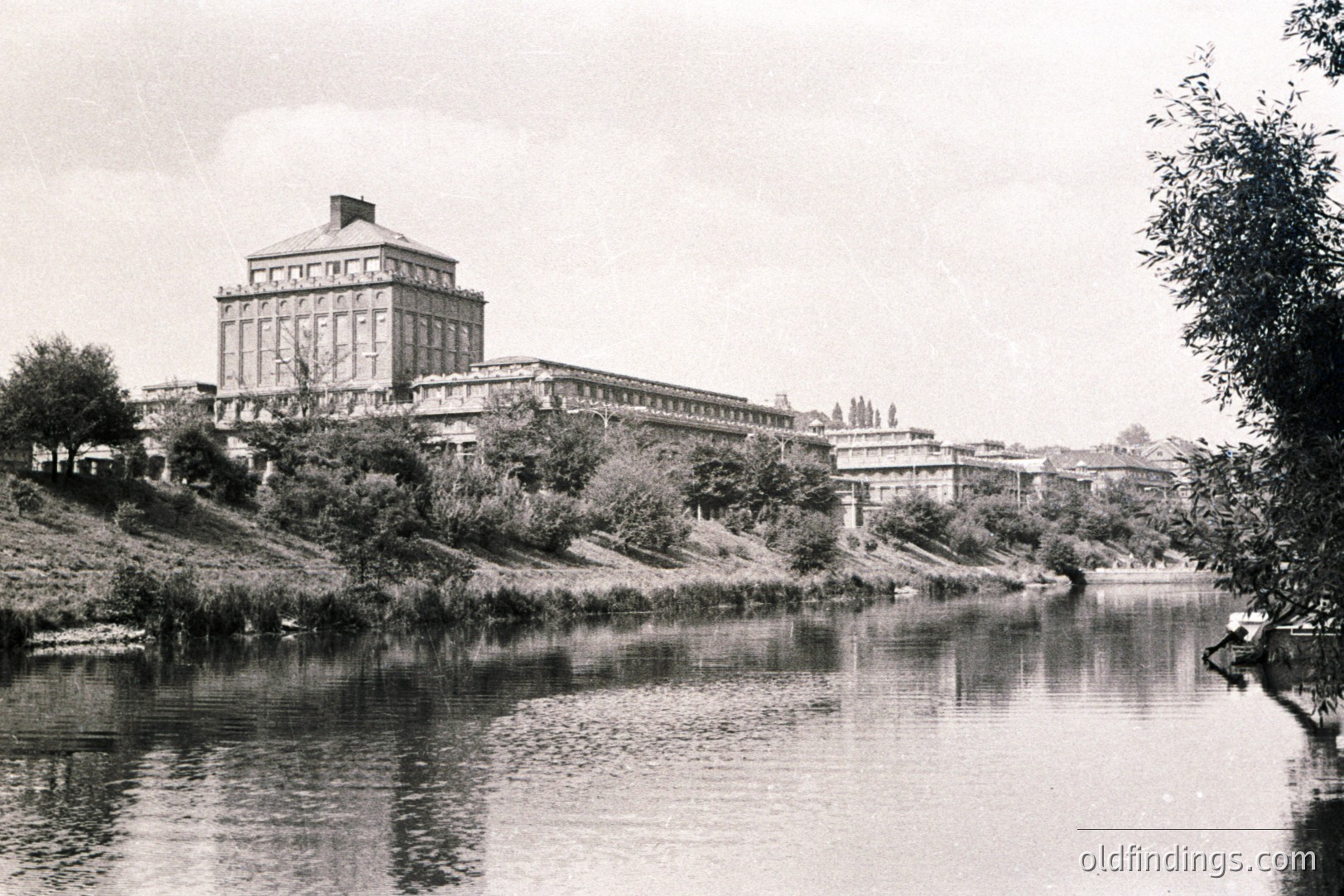 Grand, neoclassical building complex sits atop a grassy bank overlooking a river. Multiple wings extend from a central, prominent tower. Likely a public institution or university. Architectural style suggests mid-20th century, possibly 1950s-1970s. Location uncertain, but appears European.