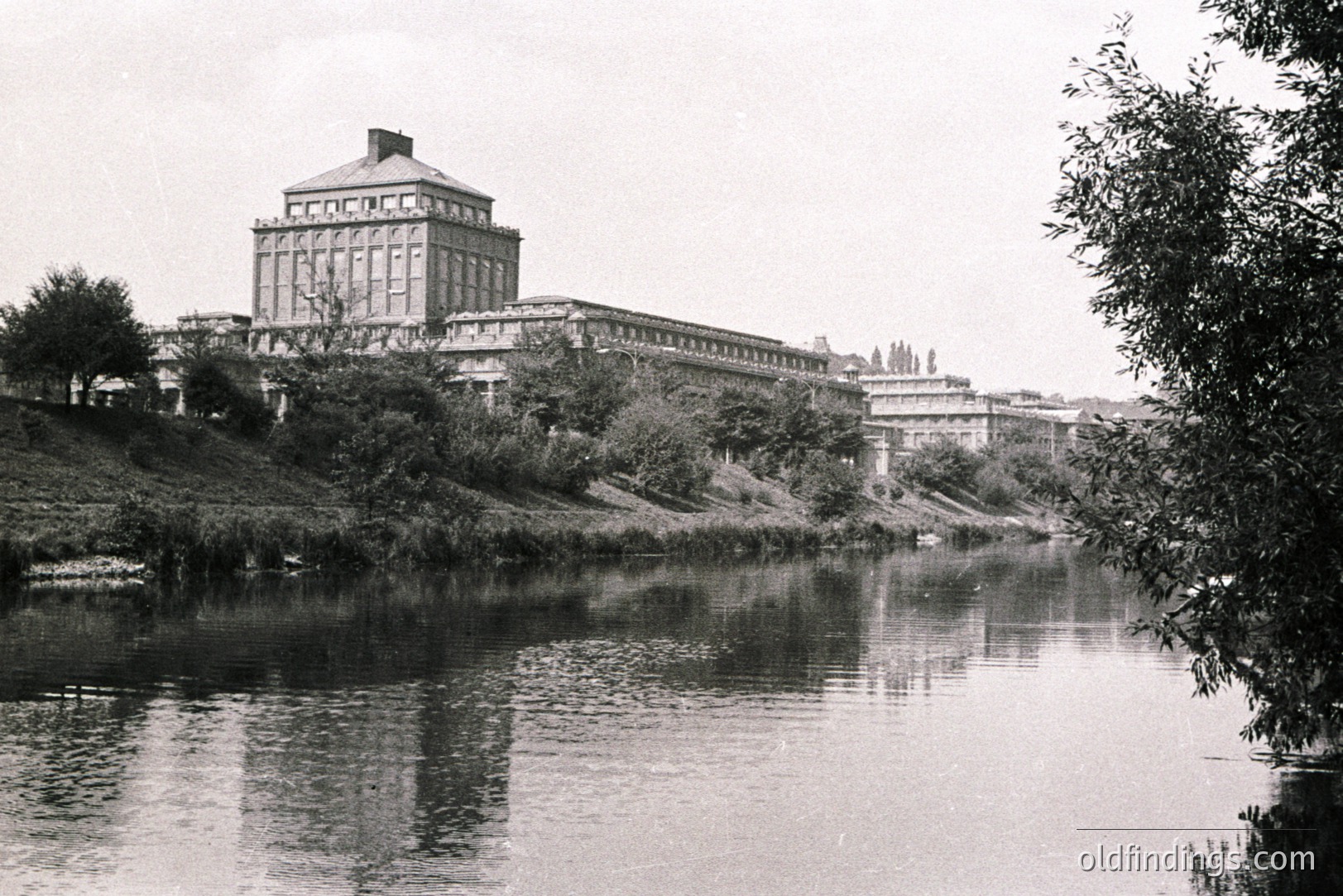 A grand, neoclassical building with a central cupola dominates the scene, situated on a raised bank overlooking a wide river or lake. Likely a public institution or university, showcasing elaborate stonework and symmetrical design. Appears to be a vintage view, possibly from the early to mid-20th century. Architectural reference value.