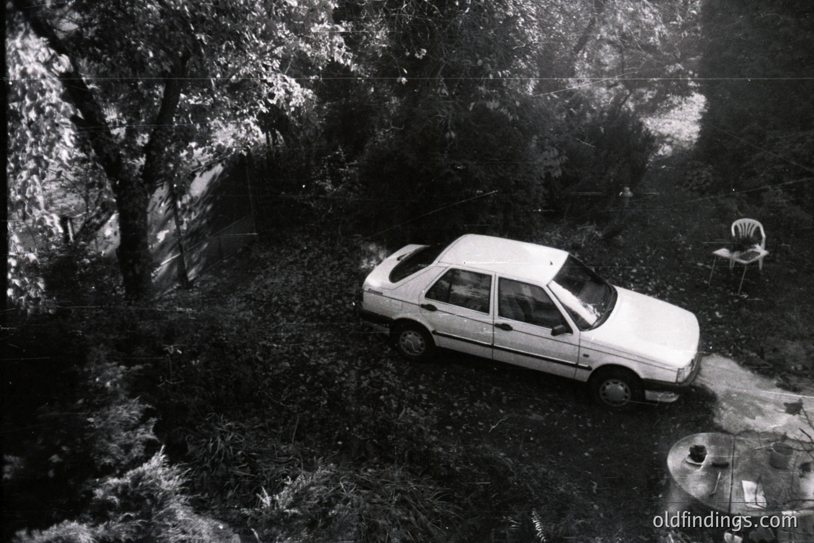 A high-angle, black-and-white shot captures a white sedan parked on an overgrown, sloping property. A patio set with a table and chairs sits nearby, suggesting a domestic scene. The vegetation is dense, indicating a rural or semi-rural location. Likely mid-1980s based on the vehicle’s design. Potential for design reference or automotive archival use.