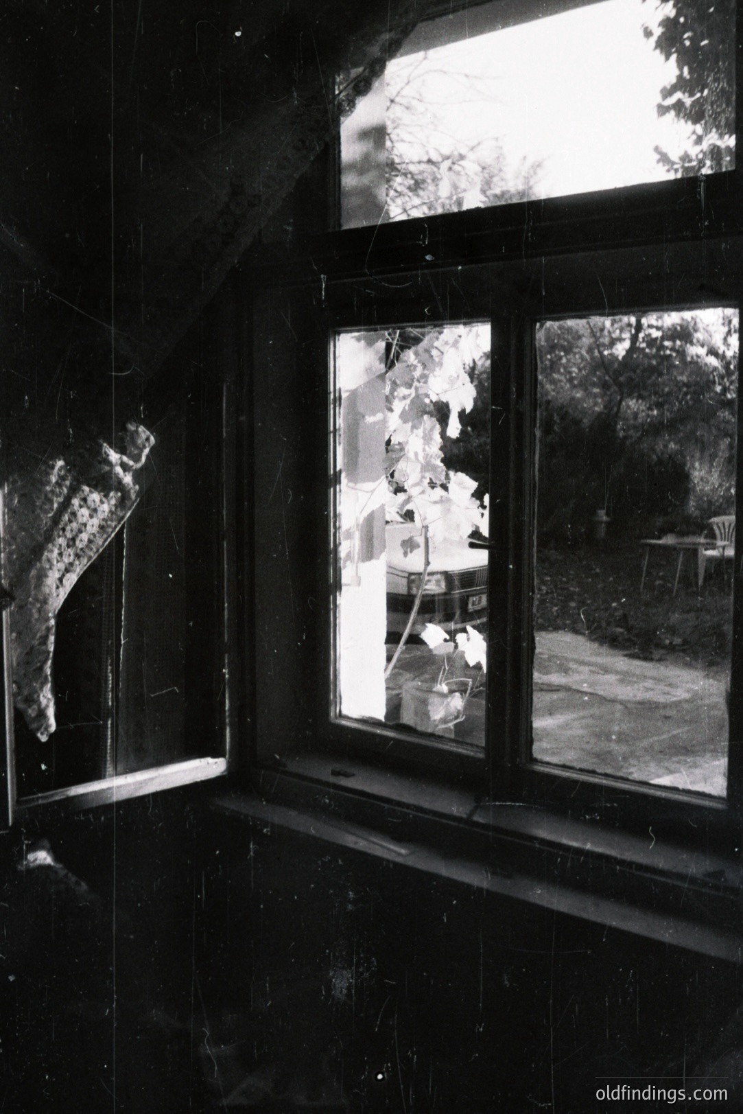 A vintage black and white photograph captures a view through a window, showing a garden scene. A loosely knit shawl hangs on a hook within the room, contrasting with the bright outdoor light. A weathered car sits in the yard amidst foliage and a patio area. Likely 1960s-70s, evocative of domestic life.