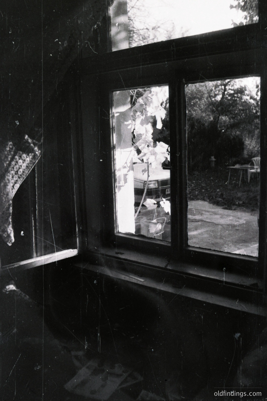 Looking through a large window framing a lush garden scene. Visible are trees, foliage, outdoor furniture, and a parked car beyond. The interior shows signs of age with visible dust and scratches on the window surface, a slightly faded textile curtain. Appears to be a mid-century home.