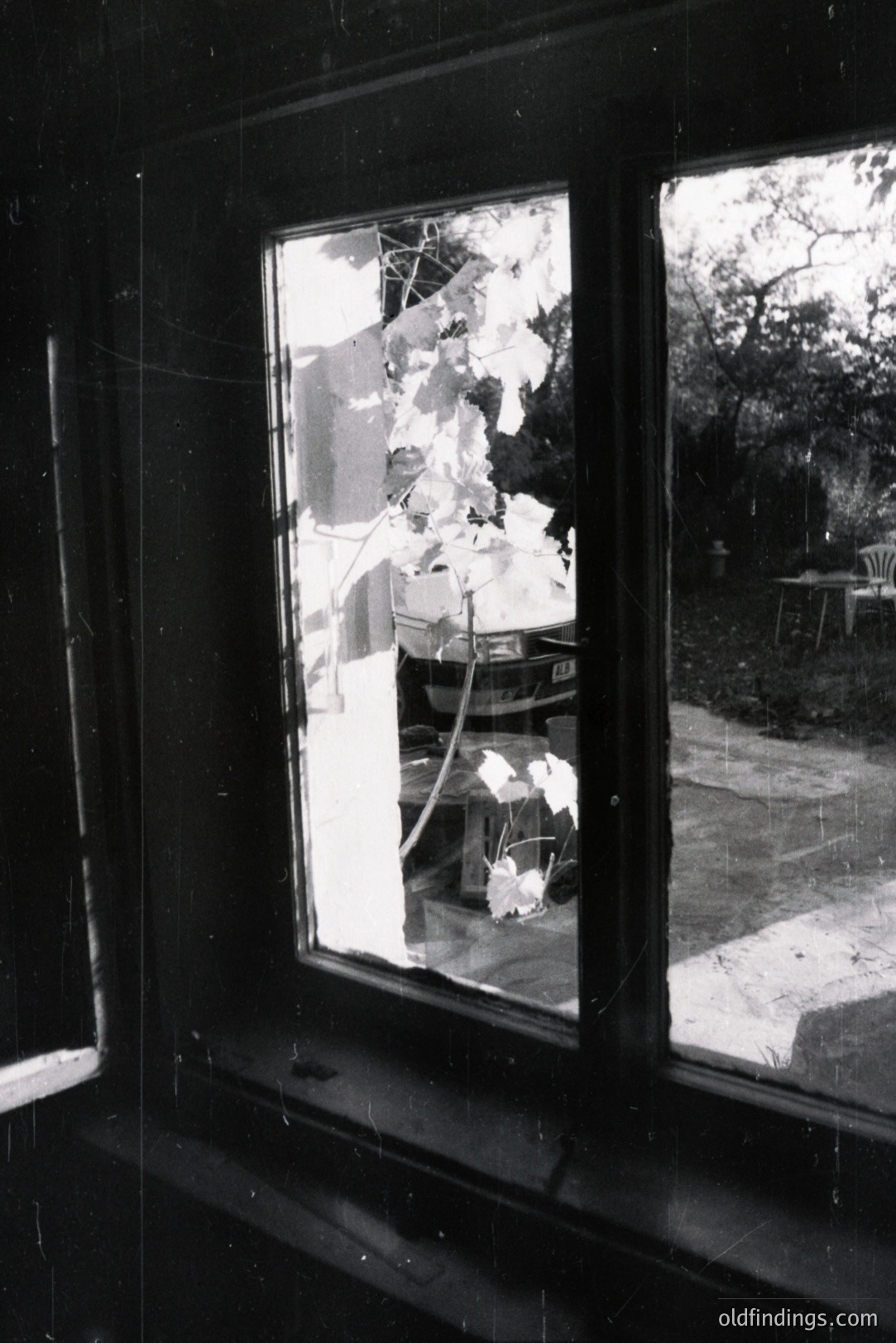Cracked window frame captures a blurred exterior scene: a car, foliage, and patio furniture. The damage suggests impact or stress. Likely mid-20th century, possibly post-war era. Evokes themes of disruption and fragility. Suitable for design referencing decay.