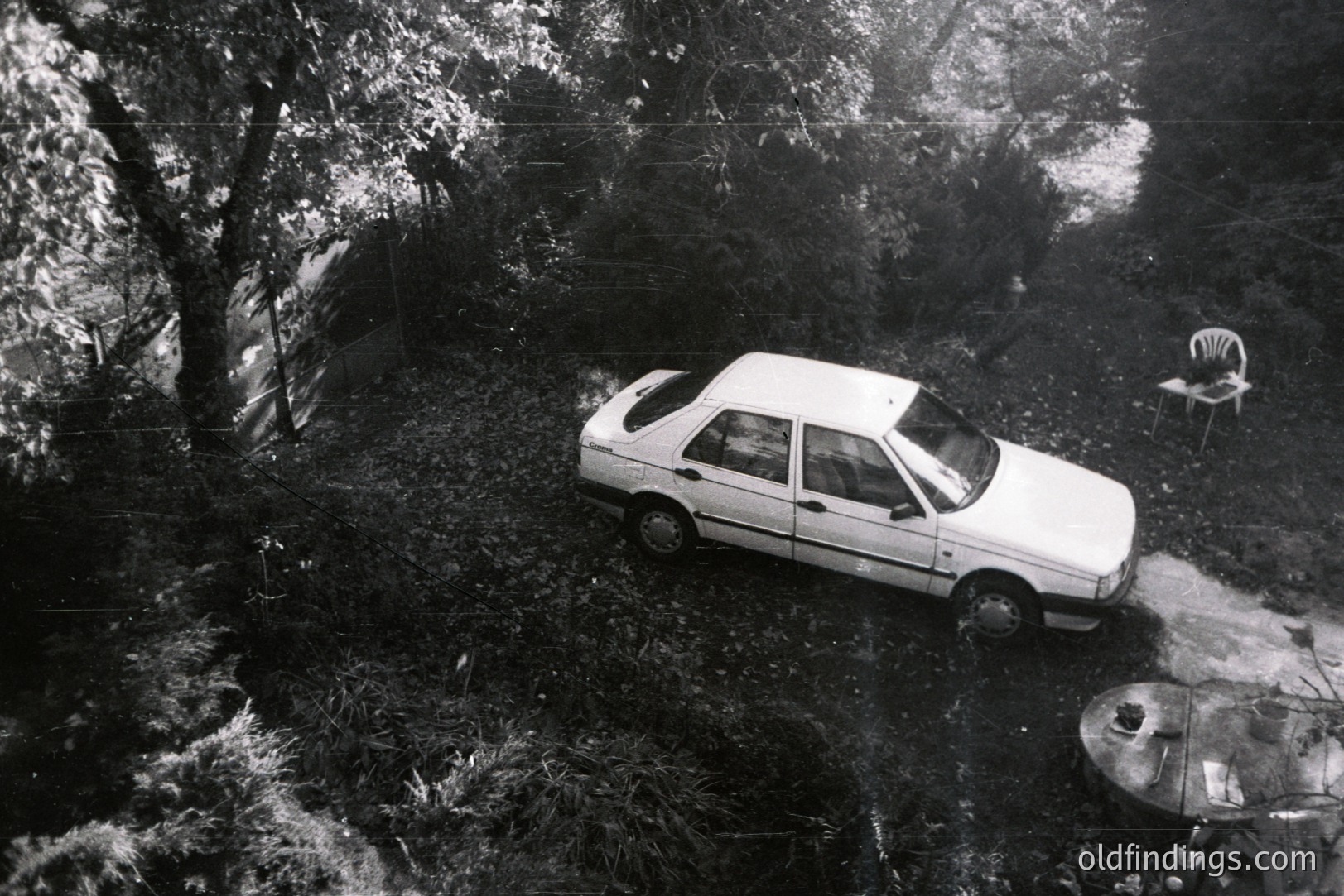 A white sedan is stranded on a patch of bare earth surrounded by dense foliage and overgrown shrubbery. A table and chairs suggest a disrupted outdoor dining area. Appears to be an aerial or high-angle perspective. Likely 1980s, possibly Eastern European setting. Unusual, evocative scene.