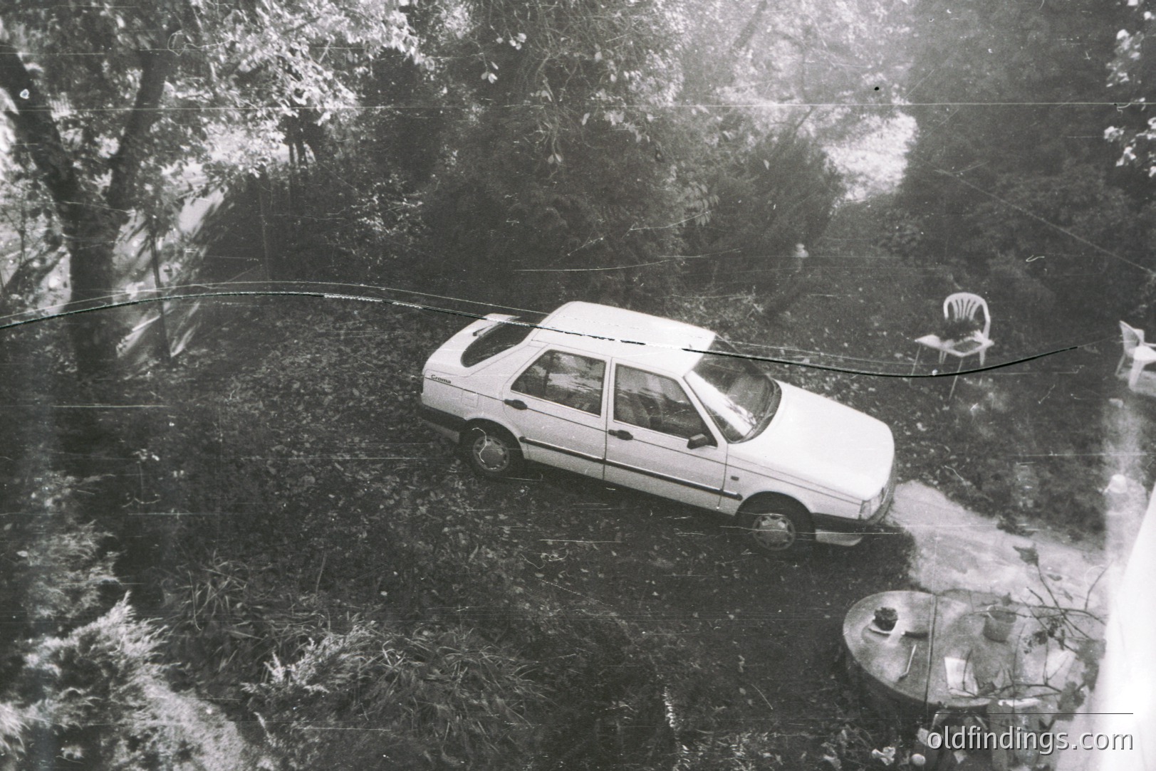 High-angle, monochrome image depicts a white sedan parked on a dirt path through a densely wooded area. A round table with dishes sits nearby, and a single chair rests on the ground. Visible damage and scratches on the photograph suggest age and use. Likely 1980s.