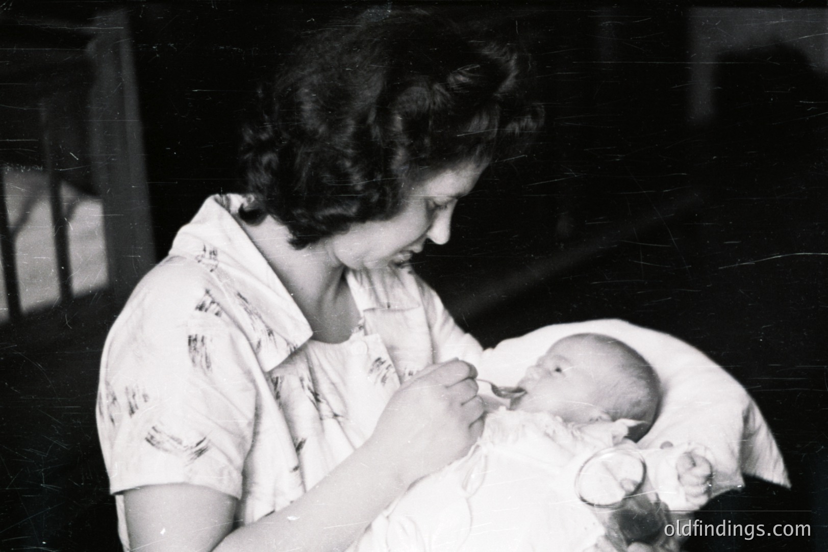 A woman in a short-sleeved, button-down gown gently cradles a sleeping infant in a hospital setting. Strong directional lighting illuminates the pair. Likely a post-natal photograph; reflective of mid-20th century family moments. Paper texture and image grain indicate age. Minimal background detail.