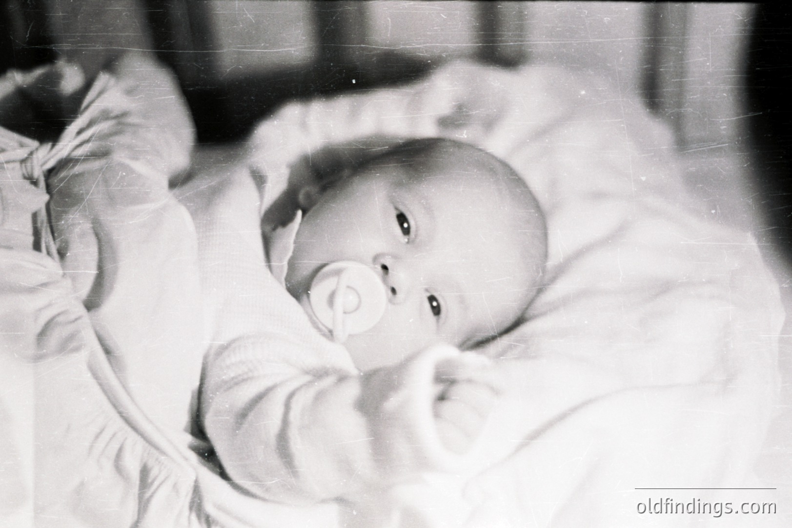 Infant rests on textured bedding, holding a pacifier. Close-up captures a direct gaze, suggesting a candid moment. Likely a family snapshot, exhibiting signs of age with visible film grain and surface scratches. Estimated 1950s-1960s.
