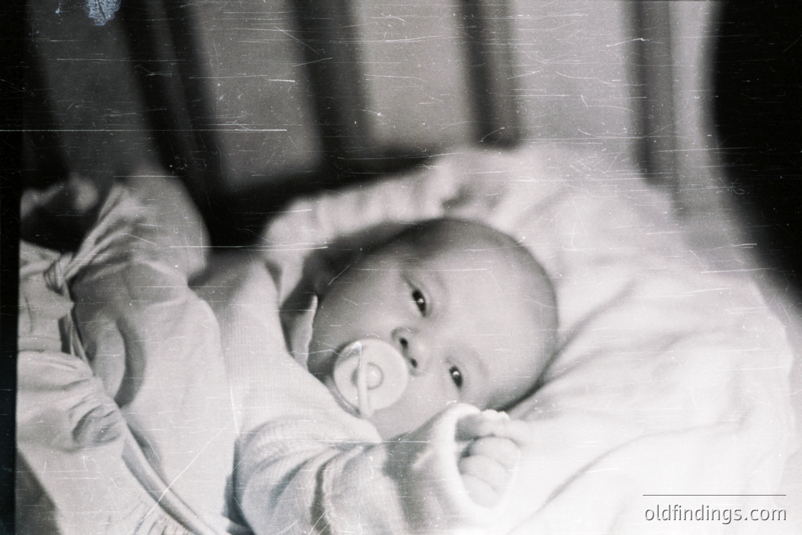 Black and white image of an infant lying on a surface, seemingly in a crib or bed. The baby wears a light-colored garment and has a pacifier in its mouth, looking directly at the camera. Visible grain and scratches indicate age. Likely a family snapshot.