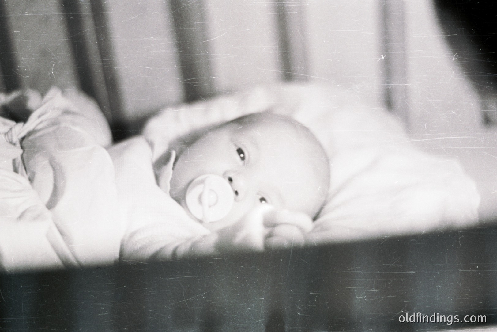 A baby lies in a crib, clutching a pacifier. The monochrome image suggests a candid, personal snapshot, likely from the mid-20th century. Visible grain and scratches are characteristic of film photography from the era. This would be valuable for archival or historical stock imagery.