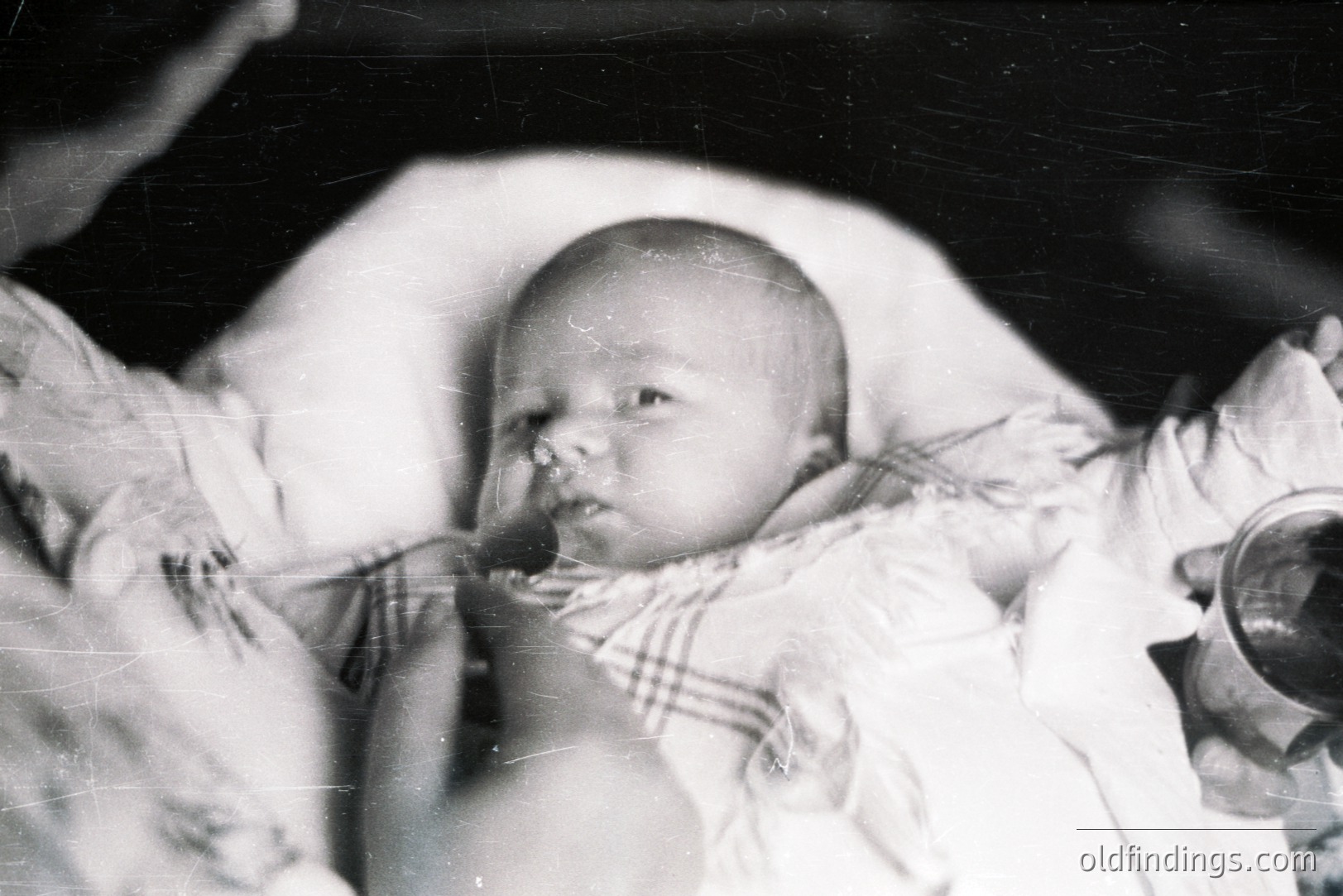 Close-up, black & white portrait of an infant cradled in arms. Baby wears a delicate, ruffled dress. Grainy texture & visible scratches suggest age. Likely a family snapshot, possibly 1930s-1950s. Potential for historical research/genealogy applications.