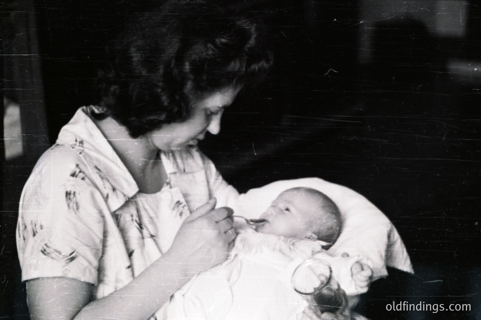 A young woman in a floral print dress tenderly feeds a baby from a bottle. The image, likely a candid moment, displays a close-up view emphasizing a maternal bond. Likely a domestic scene from the 1950s or 1960s. Black and white print shows age and wear. Suitable for historical family research or design references.