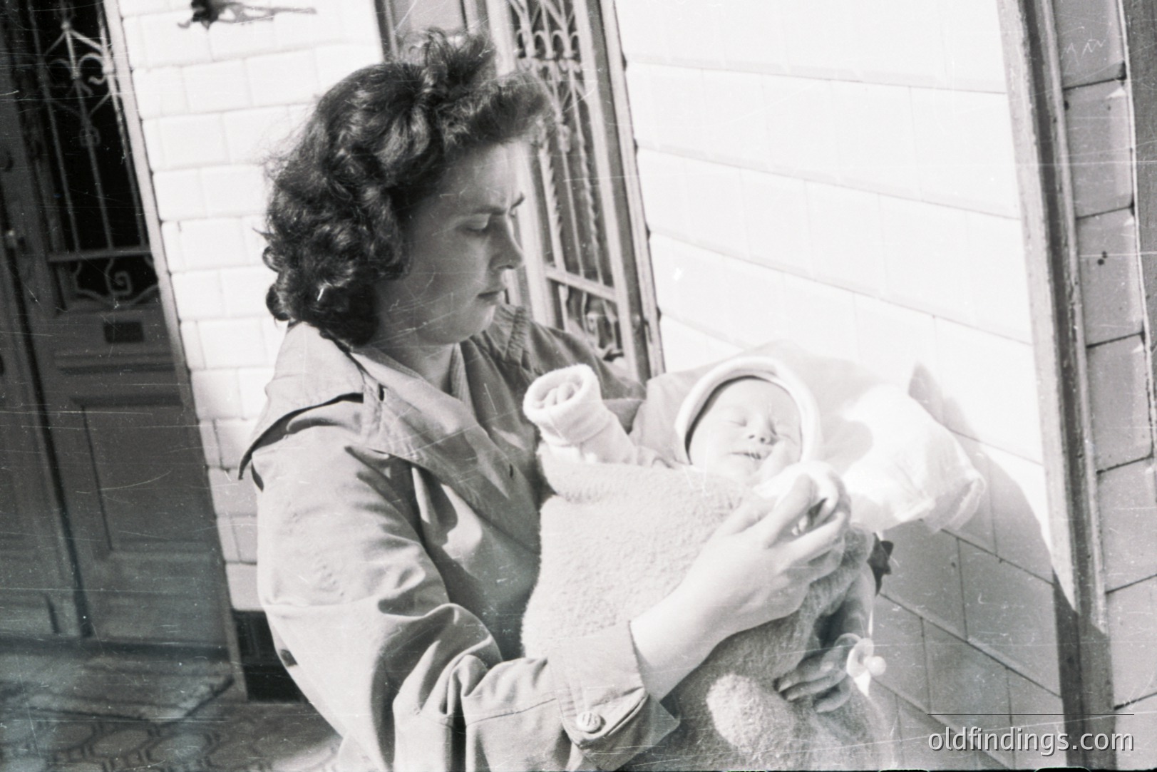 A woman, possibly a mother, holds a swaddled infant outside a building, likely a home. Architectural details suggest an early to mid-20th century setting. The woman wears a tailored coat, a style common in the 1940s or 1950s. A simple, domestic scene with an understated, classic aesthetic.