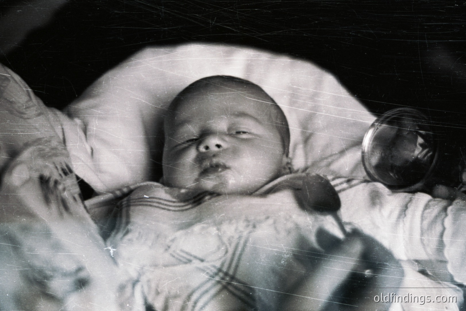 A monochrome portrait shows a peacefully sleeping baby swaddled in a striped blanket. The image exhibits visible grain and scratches typical of older photographic prints, suggesting age. A partial view of a vintage camera appears in the upper right corner. Likely a personal snapshot, potentially from the mid-20th century.