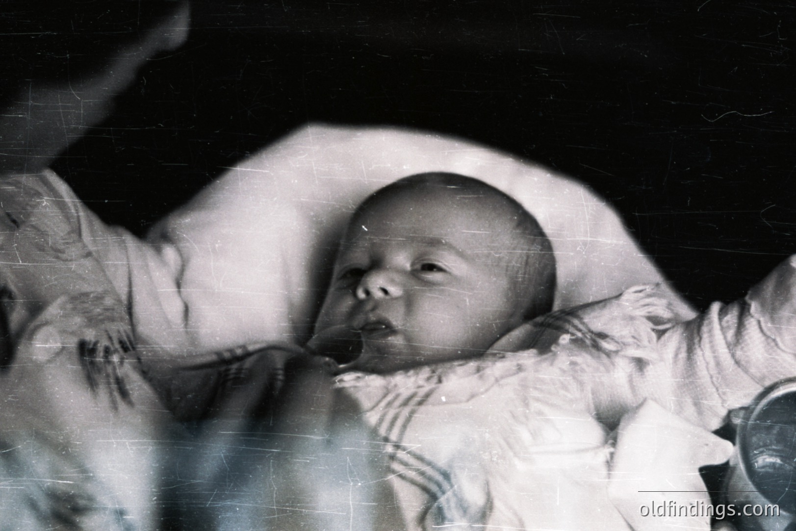 A monochrome portrait of an infant in a high-collared, lace-trimmed garment. The subject appears to be mid-expression, possibly reaching for something beyond the frame. Likely a family snapshot, dating from the mid-20th century. Visible film grain and surface scratches suggest age and possible archival value.
