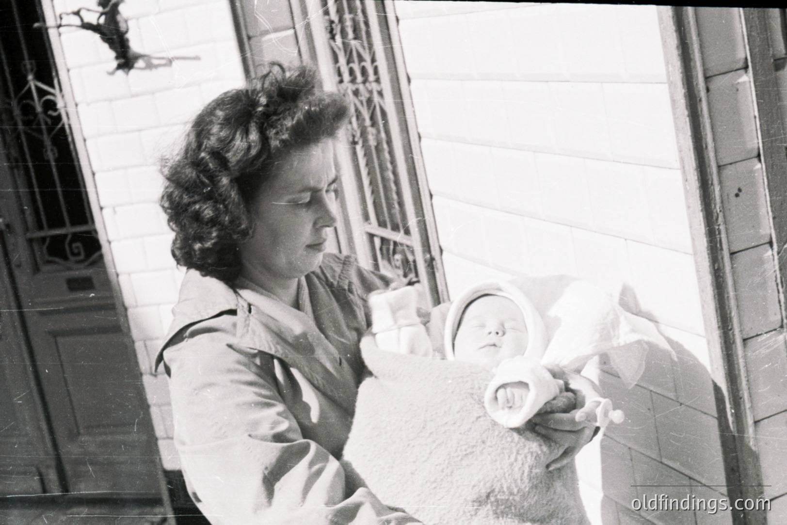 A young woman holds a swaddled baby on a porch with ornate wrought-iron details. Likely taken in the 1940s or 50s, it portrays a domestic scene with vintage clothing styles and a formal portrait aesthetic. The architectural details suggest a well-maintained, possibly older home. A poignant moment of maternal care and early childhood.