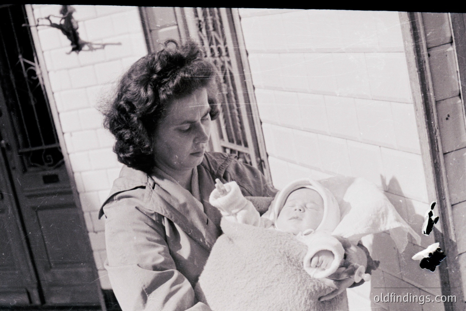 A young woman holds a bundled infant, likely a newborn, in a brick-walled exterior. Visible architectural details suggest a residential setting. The woman’s hairstyle and clothing indicate a mid-20th-century (1940s-1960s) timeframe. Image shows intimate family moment. Historical documentation of early parenthood.