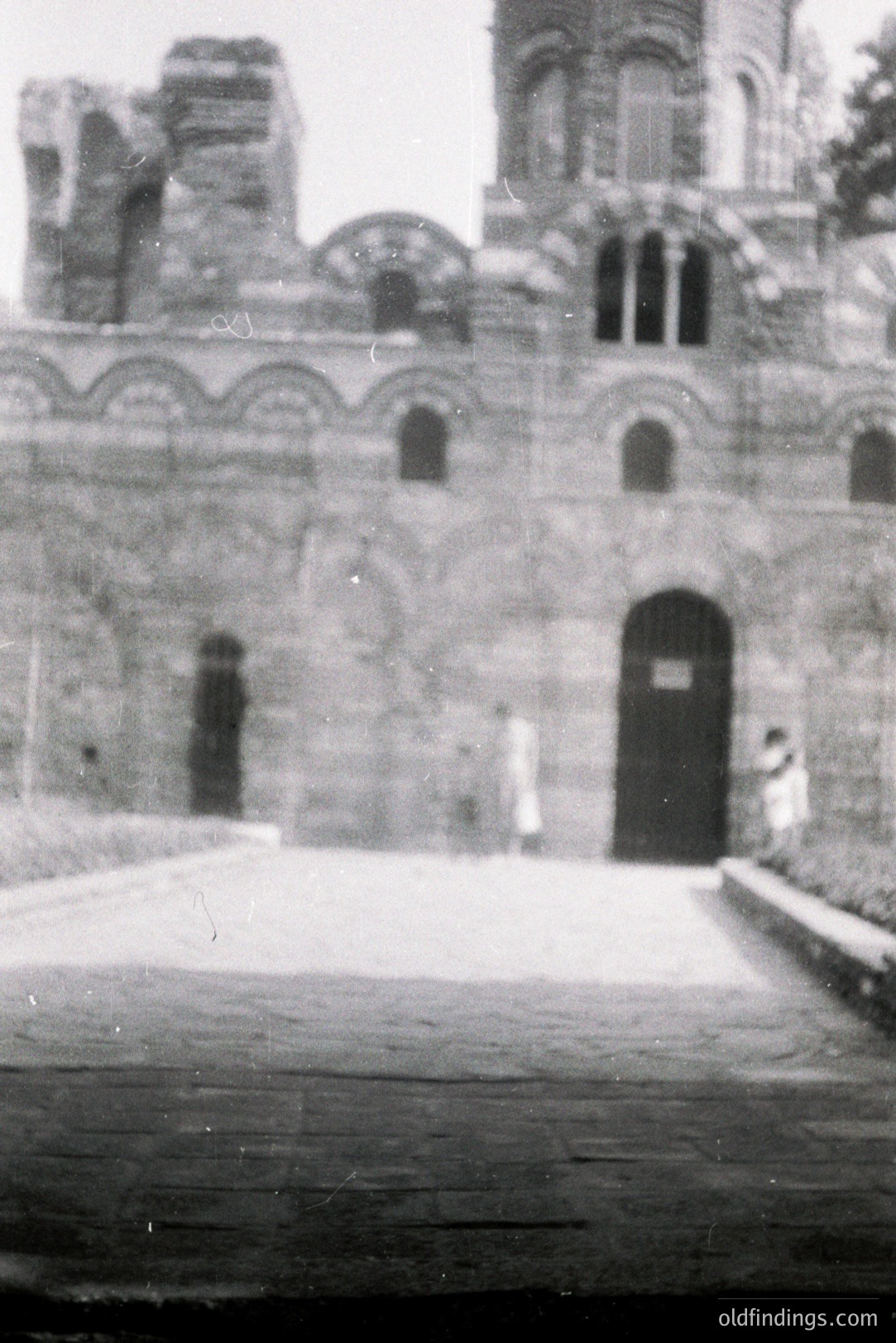 Monochrome photograph depicting the facade of a stone building, likely a church or monastery. Arched windows punctuate the stonework. Two figures are faintly visible near the entrance. Appears to be a historic building, potentially from the 19th or early 20th century.