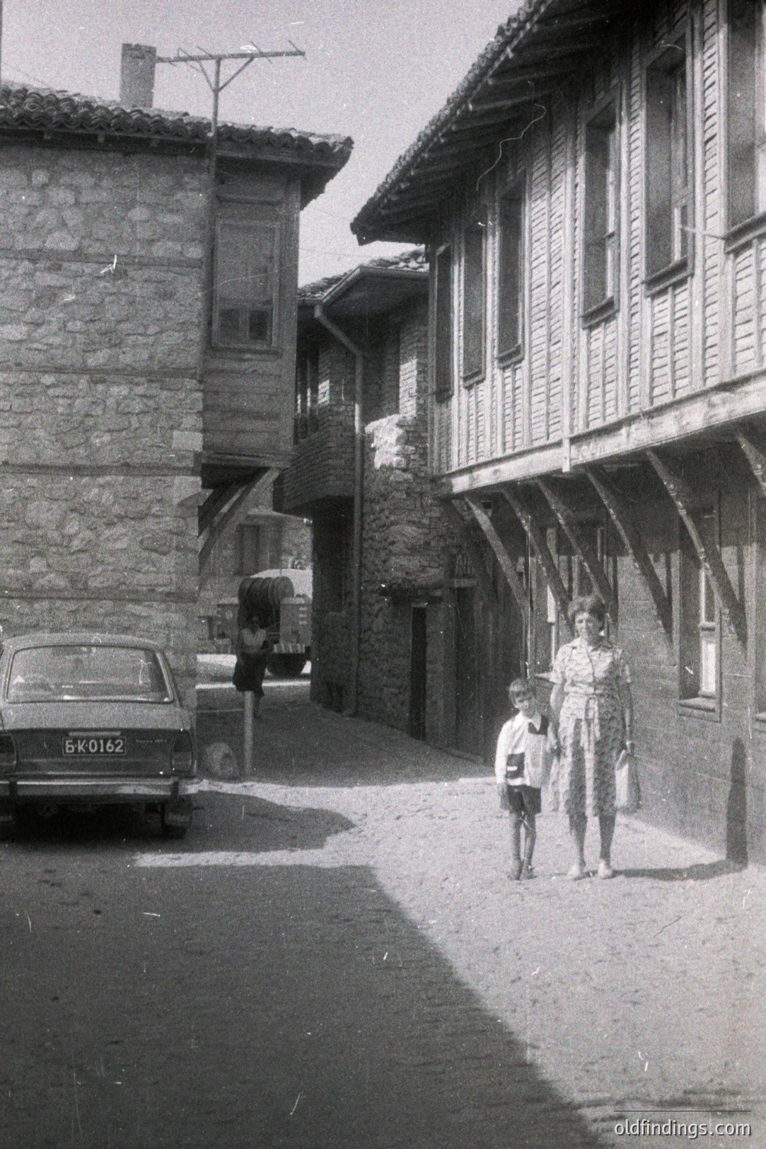 A young boy and woman walk down a narrow, cobblestone street lined with traditional timber-framed buildings. A car (license plate "БK0162") is parked along the side. Likely Bulgaria, capturing architecture and daily life, possibly 1970s. Valuable for cultural studies & design.