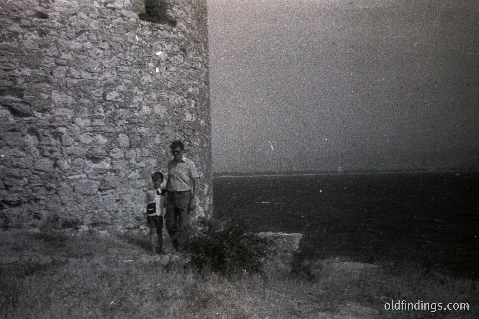 Two figures stand near a rough-hewn stone wall overlooking the sea. The wall appears to be part of an older structure, likely coastal fortifications. The subjects, a young boy and older individual, are casually dressed. Location possibly coastal Bulgaria, Varna area. Likely 1960s or 70s amateur photography. Shows architectural detail and potential tourist activity.