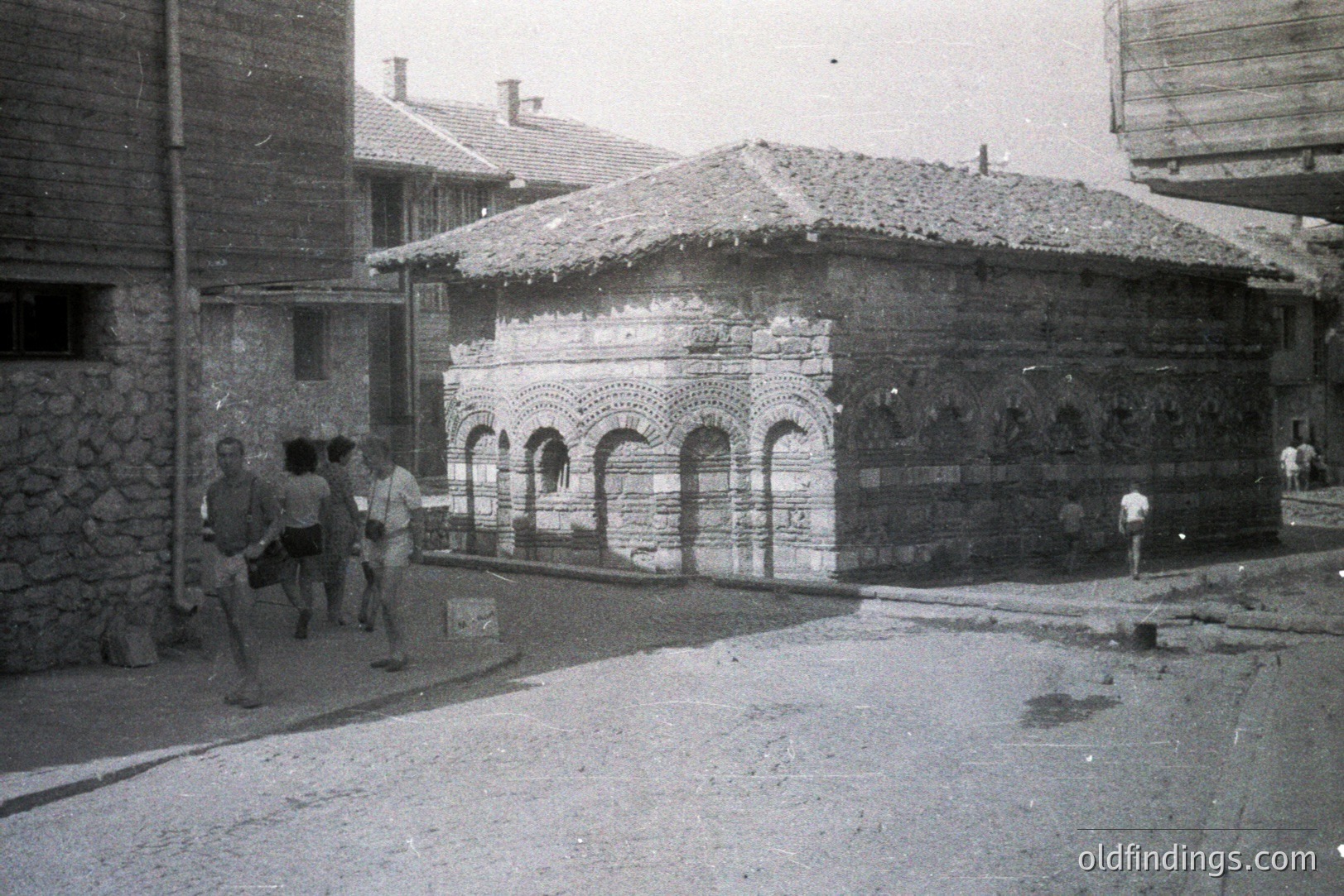 A unique, compact building with arched brickwork and a low, tiled roof stands on a dusty, cobbled street. Three men in swimwear walk past. The architecture suggests a public bathhouse or similar utility structure, likely from the 1960s. Appears to be Bulgaria, potentially Varna.