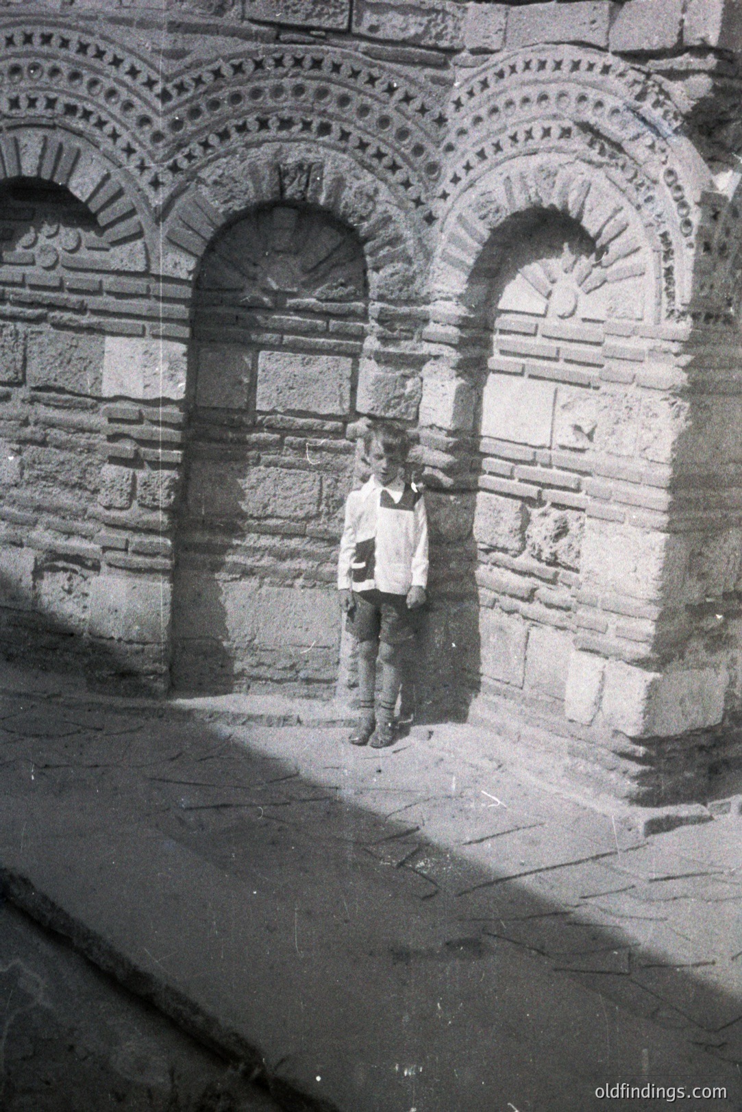 A young boy stands beneath a series of arched stone openings. The structure’s exterior features detailed relief carvings around the arches. A faded inscription is visible on the wall. Likely early-mid 20th century, judging by the boy’s clothing and photographic style. Location unknown. Potential for architectural study.