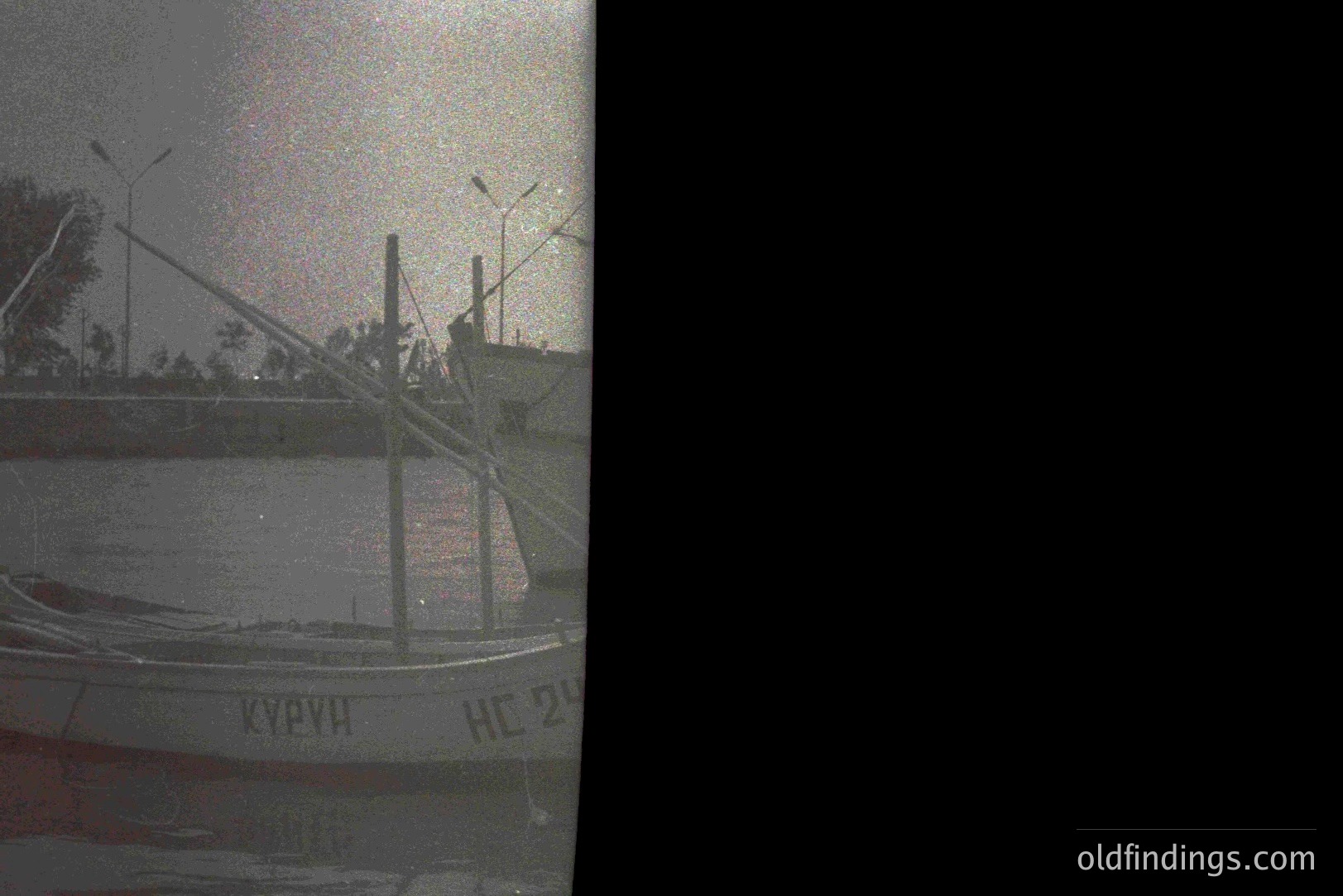 A photograph shows a weathered boat with Cyrillic script ("Курун" and "HC 24") on its side, reflecting a landscape. Tall utility poles with overhead wires suggest a coastal industrial area. Possible location: Black Sea region. Appears to be from the mid-20th century. Image exhibits significant film grain.