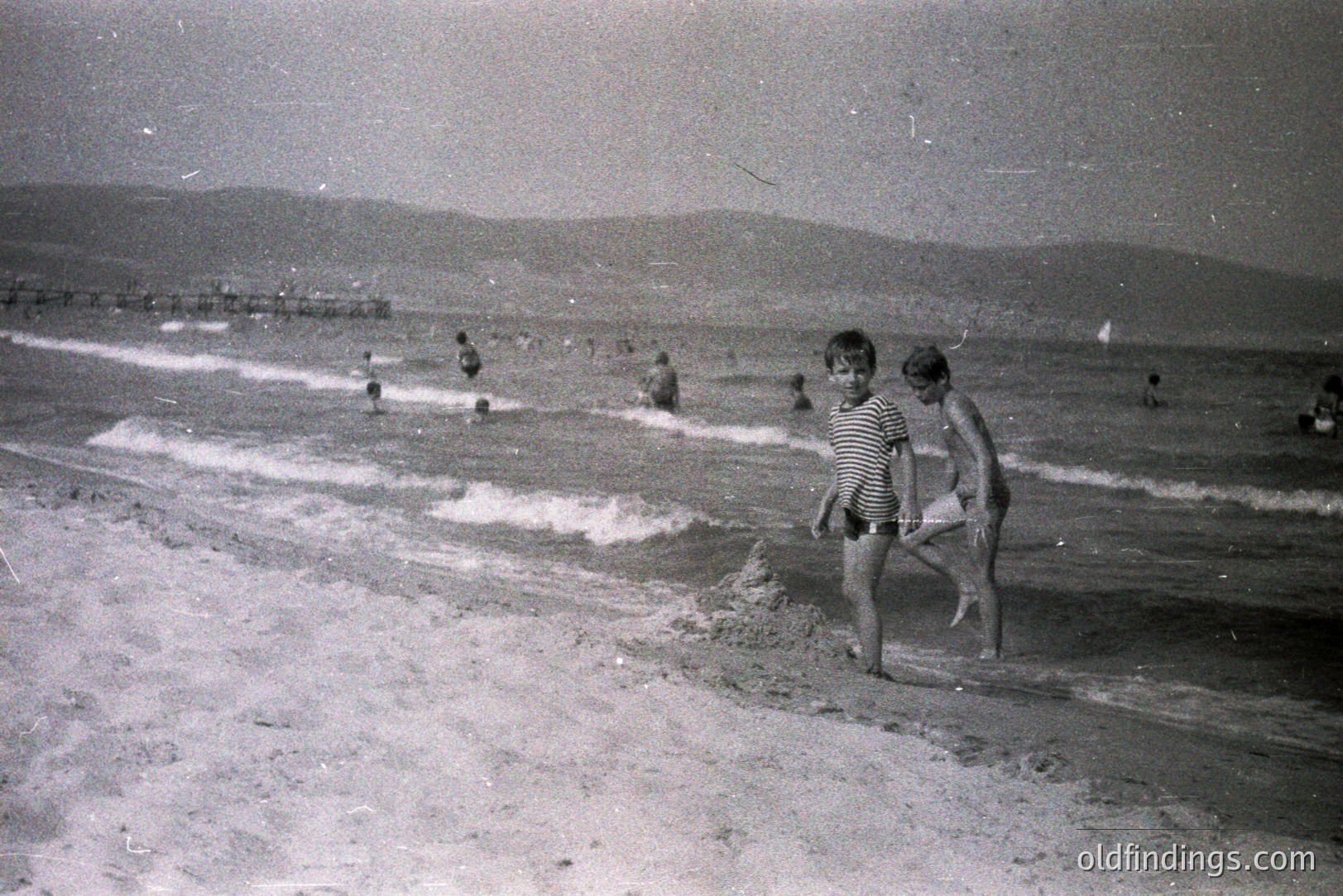 Two young boys, one striped shirt, wade along a sandy beach with gentle waves. Figures are scattered in the background. A distant pier and low hills suggest a seaside location, possibly Eastern Europe. Likely 1960s-1970s amateur photography. Evokes nostalgic summer memories.