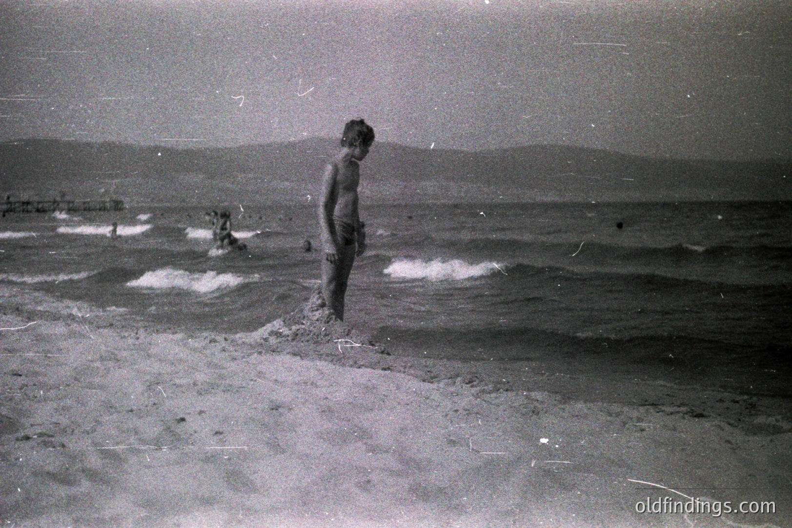 A young, barefoot boy stands on a sandy beach facing the sea, possibly contemplating the waves. Distant figures and a structure are visible along the shoreline. The grainy image suggests a vintage film photograph, likely from the 1960s or 70s. Location undetermined, but coastal environment is evident.