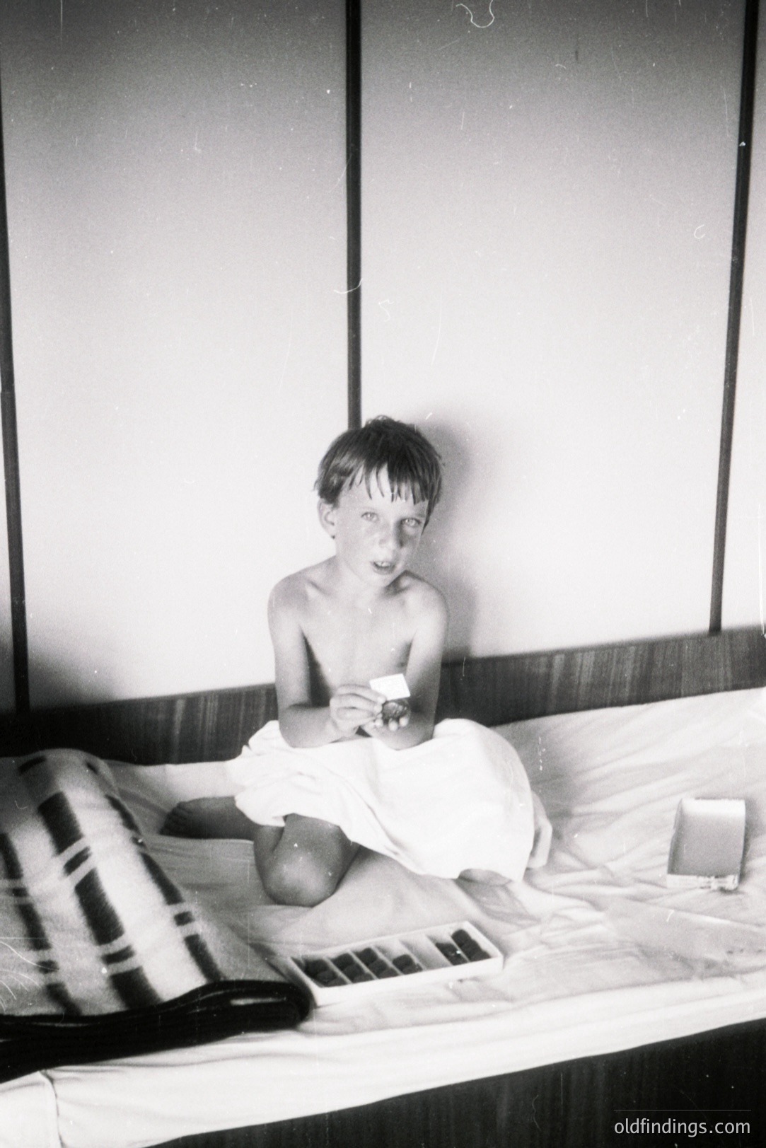A young boy, shirtless and wearing only shorts, sits on a bed holding a small rectangular object, likely a photograph. The room features paneled walls and a striped blanket. Likely a candid family snapshot, possibly from the 1960s or 70s. Commercial value: nostalgic family moments.