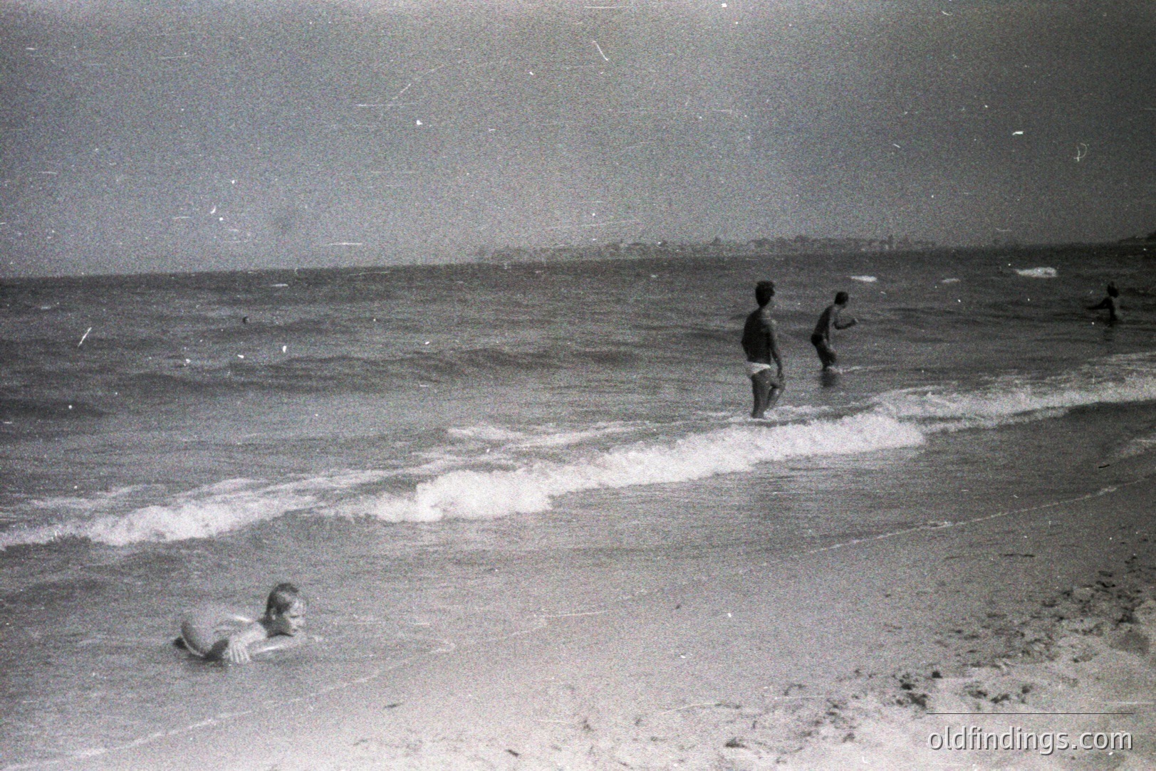 Seascape view features two boys wading in the shallows, a third person swimming, and a distant coastline. The grainy, vintage aesthetic suggests a mid-20th century snapshot. Likely a seaside resort scene, possibly Eastern Europe. The photograph holds value for documenting leisure and coastal life.