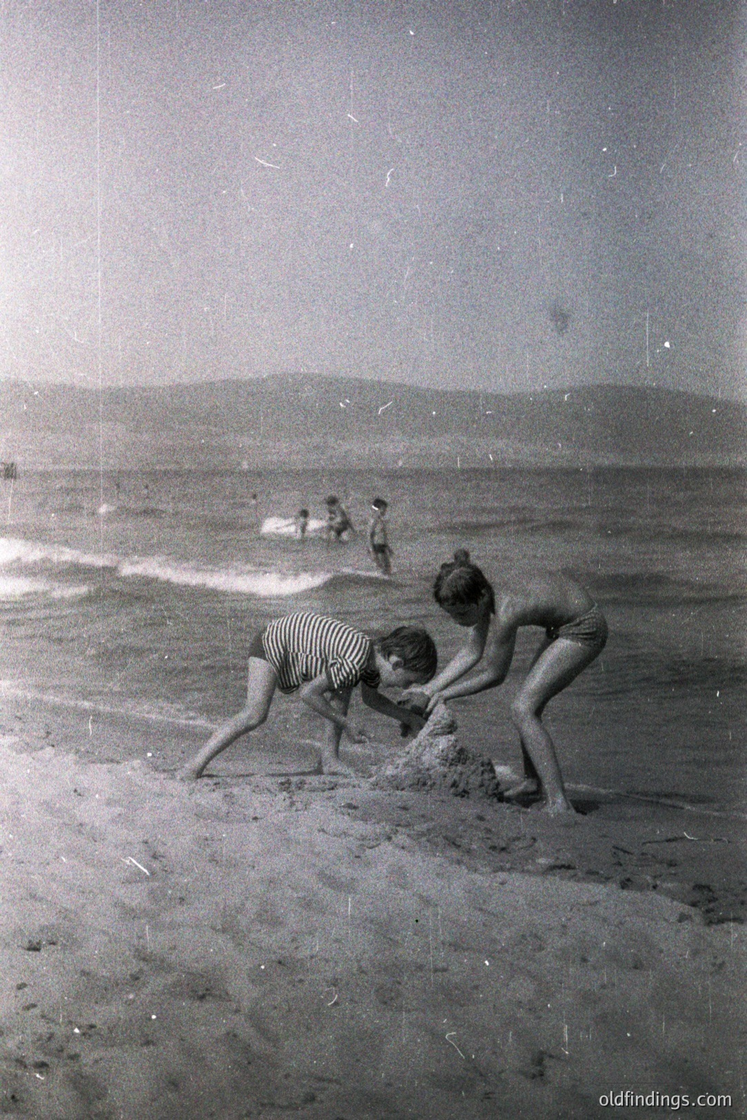 Two children building a sandcastle on a wide beach; figures in the distance. Soft focus, suggesting a candid snapshot. Likely 1960s or 70s; swimwear styles confirm this period. Coastal landscape with gentle dunes visible. Appears to be a family vacation image. Potential for historical travel/family research use.