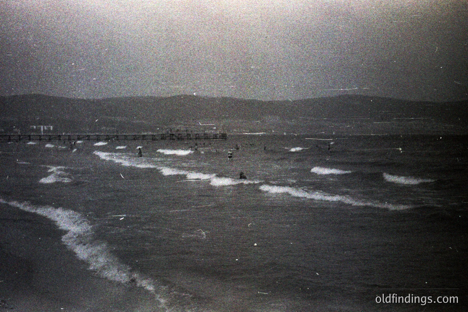 Dramatic grayscale view of a stormy seaside. Rough waves break along a wide, sandy beach under a heavy, overcast sky. A distant pier extends into the water with small figures visible along its length. Likely a tourist destination, possibly early color photography. Potential stock photo reference.