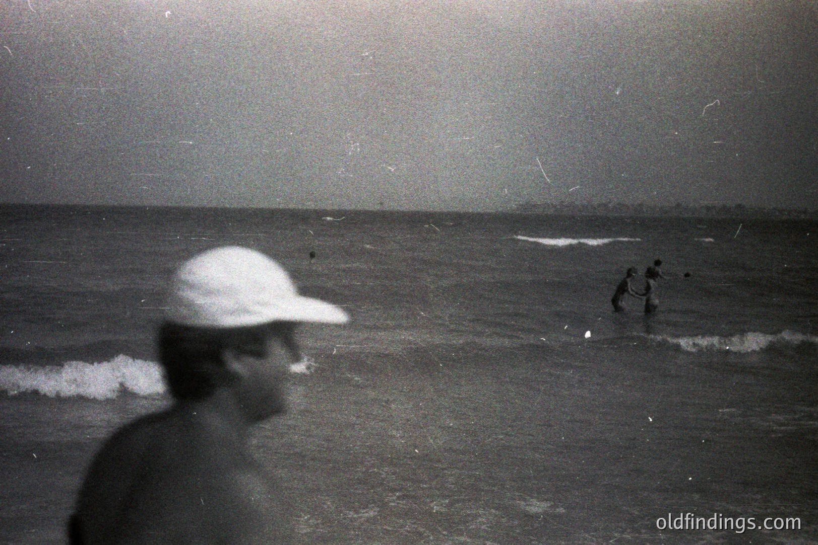 A blurry, candid monochrome captures a coastal scene. A figure in a baseball cap stands near the water’s edge, partially obscured by motion blur. Further out, two figures are seen wading in the sea. Possible 1970s aesthetic, grain evident. Evokes a nostalgic, documentary feel. Commercial value as stock imagery for coastal themes.