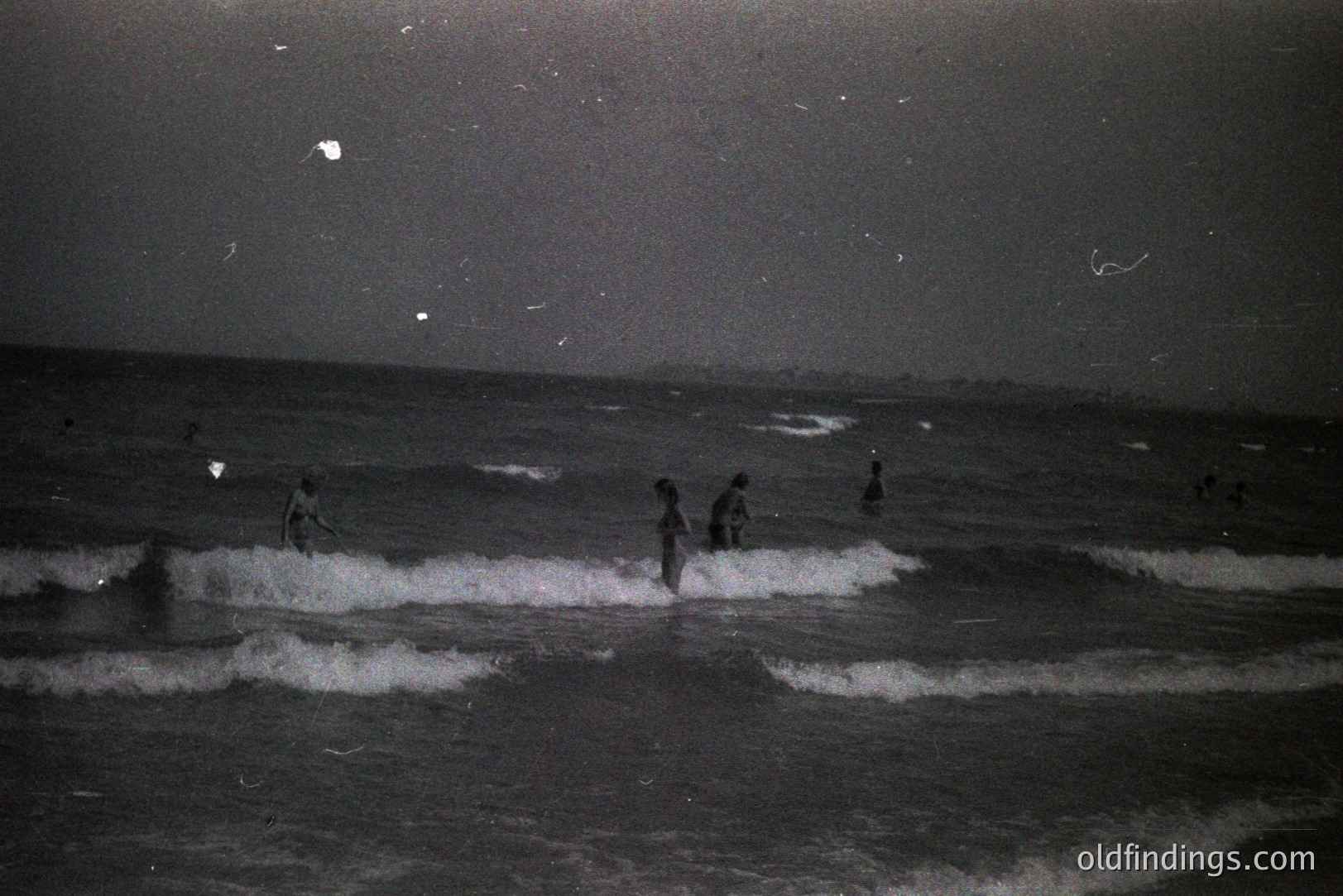 A black and white photograph depicts figures wading through breaking waves at night. The scene, likely a seaside location, shows a dark shoreline receding into the distance under a starry sky. Appears to be an archival image given grain and damage. Could be stock for atmospheric beach scenes or period research.