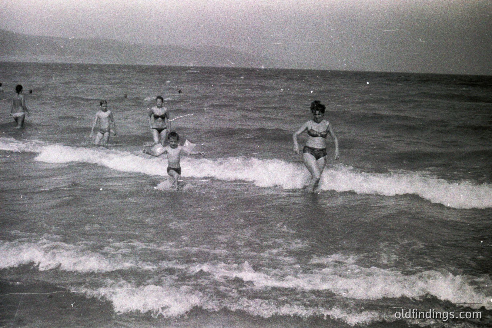 A candid moment capturing a family enjoying a day at the beach. Several figures wade into the breaking waves, a young boy in the foreground with outstretched arms. Likely a resort or vacation destination, possibly in Eastern Europe given the landscape. Appears to be from the 1960s-1970s, based on clothing and photographic style. Could be valuable for vintage travel or family portraiture stock.