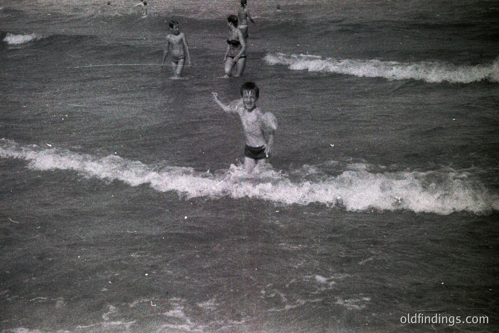 A black and white image depicts three young boys playing in the surf. The central boy, mid-stride, raises his arm in a wave. Two other boys are further out in the water. The image evokes a nostalgic, candid moment of childhood play. Likely 1960s or 70s.