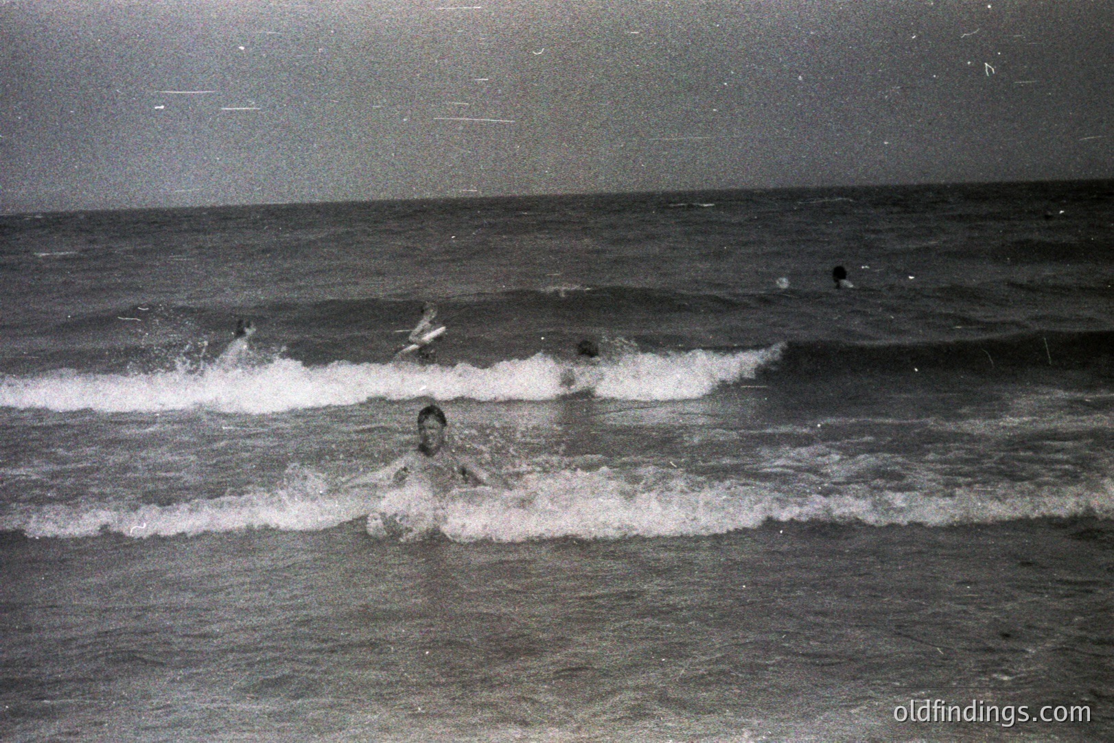 A monochrome photograph shows several figures wading through breaking waves in a coastal setting. The image captures motion and spray as the individuals navigate the water. Likely a resort or leisure scene, evoking a sense of recreation. Appears to be from the mid-20th century, possibly 1950s-1970s.