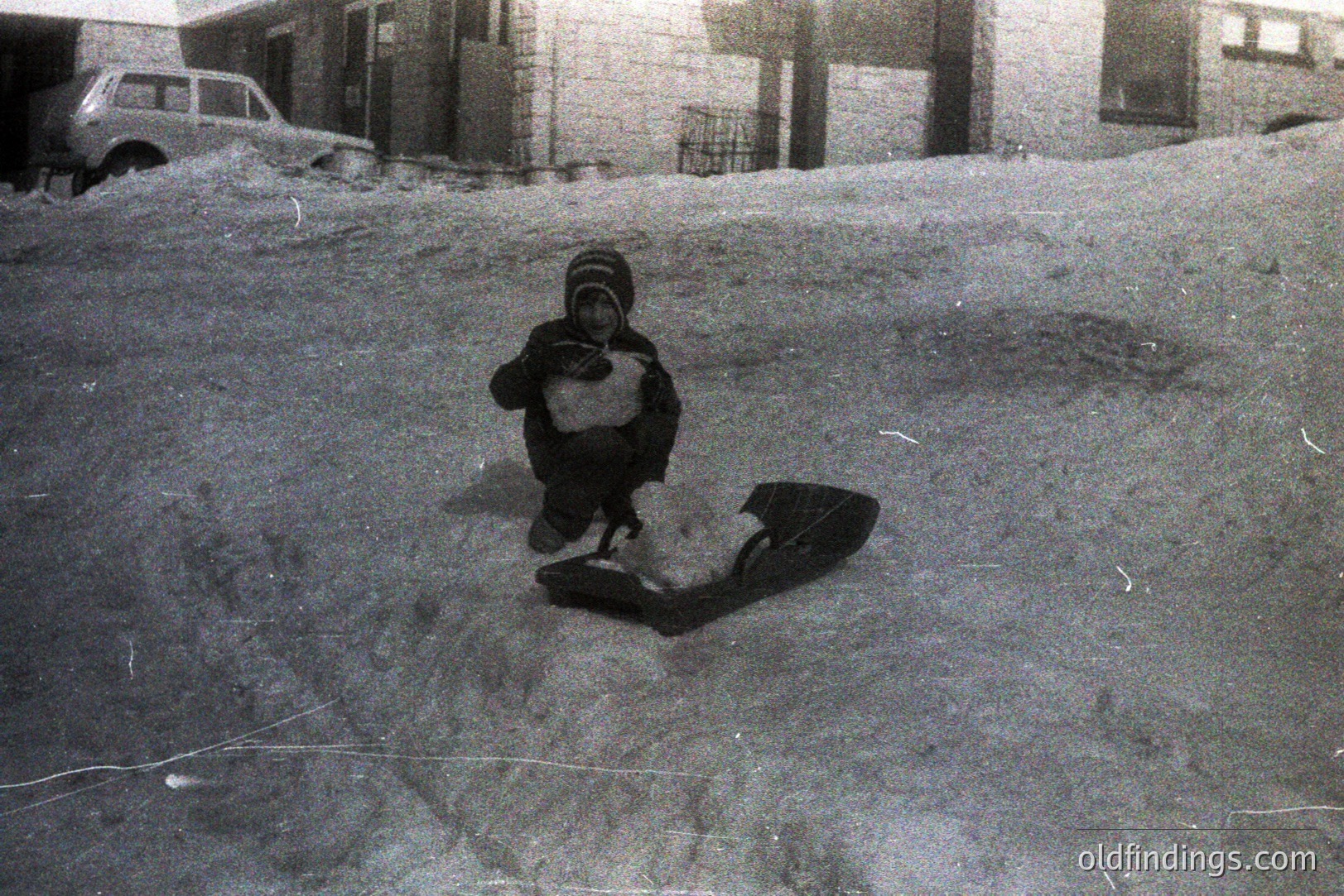 A child in winter clothing, including a hooded hat, crouches on a snow-covered slope, apparently operating a primitive sled. Architectural elements suggest a residential area with snow accumulation. Grainy texture indicates potential early photography, likely 1950s-1970s. Appears to be a candid, family moment.