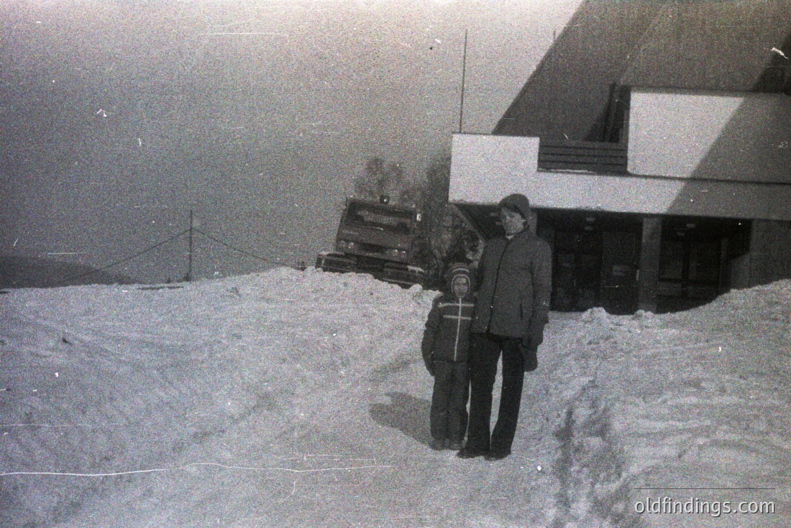 Two figures, a man and child, stand in deep snow, partially obscuring a building’s entrance. Building features a covered porch & steeply pitched roof. Appears to be a rural, possibly alpine, setting. Image exhibits grain and wear common to film photography. Likely 1960s-1970s.
