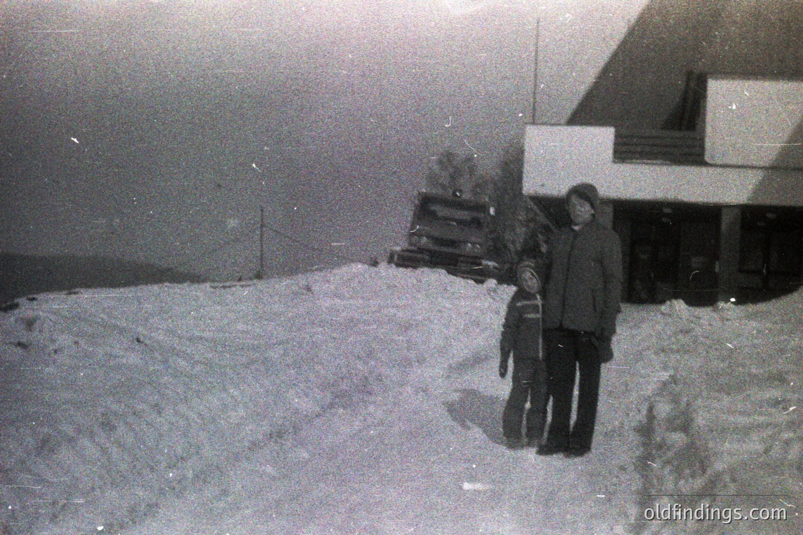 A snowy landscape with a man and child standing near a timber-framed building, likely a lodge or cabin. The grainy black and white image suggests a 1960s or 70s time period. Snow covers the ground and clings to the building’s roof and eaves. A rustic, winter scene.
