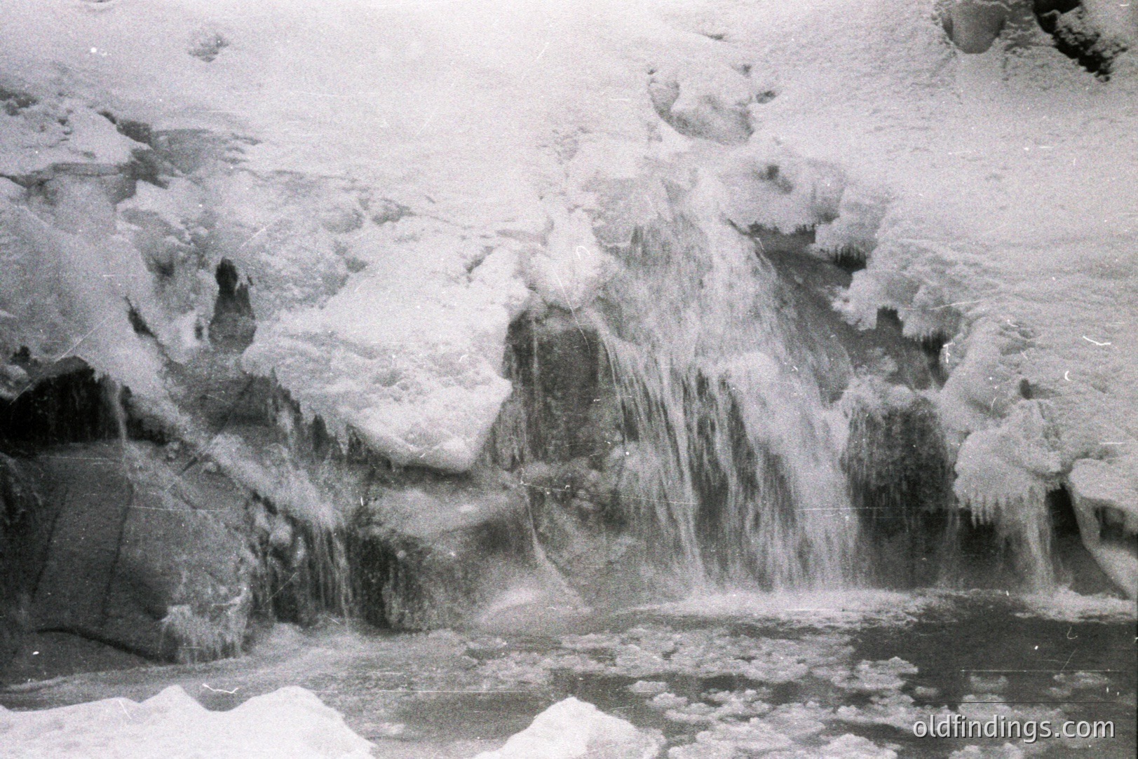 Frozen waterfall cascades over ice-encrusted rock formations. Ice structures cling to the rock face, exhibiting intricate patterns. Water pools at the base, partially frozen with ice formations. Appears to be a high-altitude, winter landscape. Likely mid-20th century based on the photographic style. Potential stock photo value for winter/ice themes.