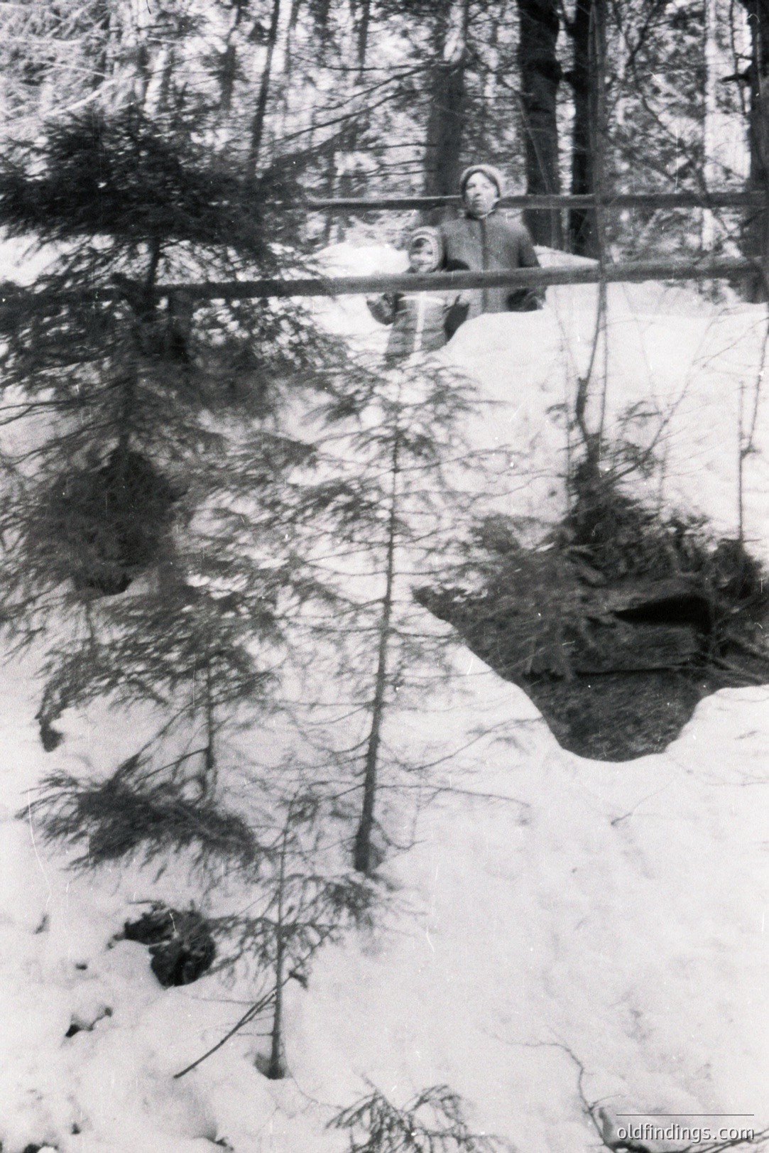 A snow-covered landscape with evergreen trees. A person in a light-colored sweater stands on a raised wooden structure, possibly a bridge or platform. The scene evokes a rural, winter setting, likely mid-20th century, judging by the photographic style. Appears to be a personal snapshot, showing natural winter beauty.