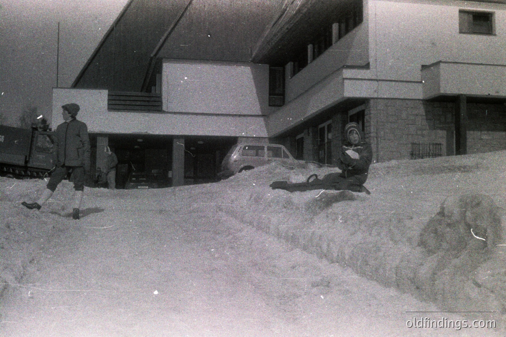 Monochromatic scene depicts a modern, concrete building with an overhanging design. A man in a hat strides across a large snow drift towards the building's entrance. An older car is partially visible near the entrance. Likely 1960s architecture.