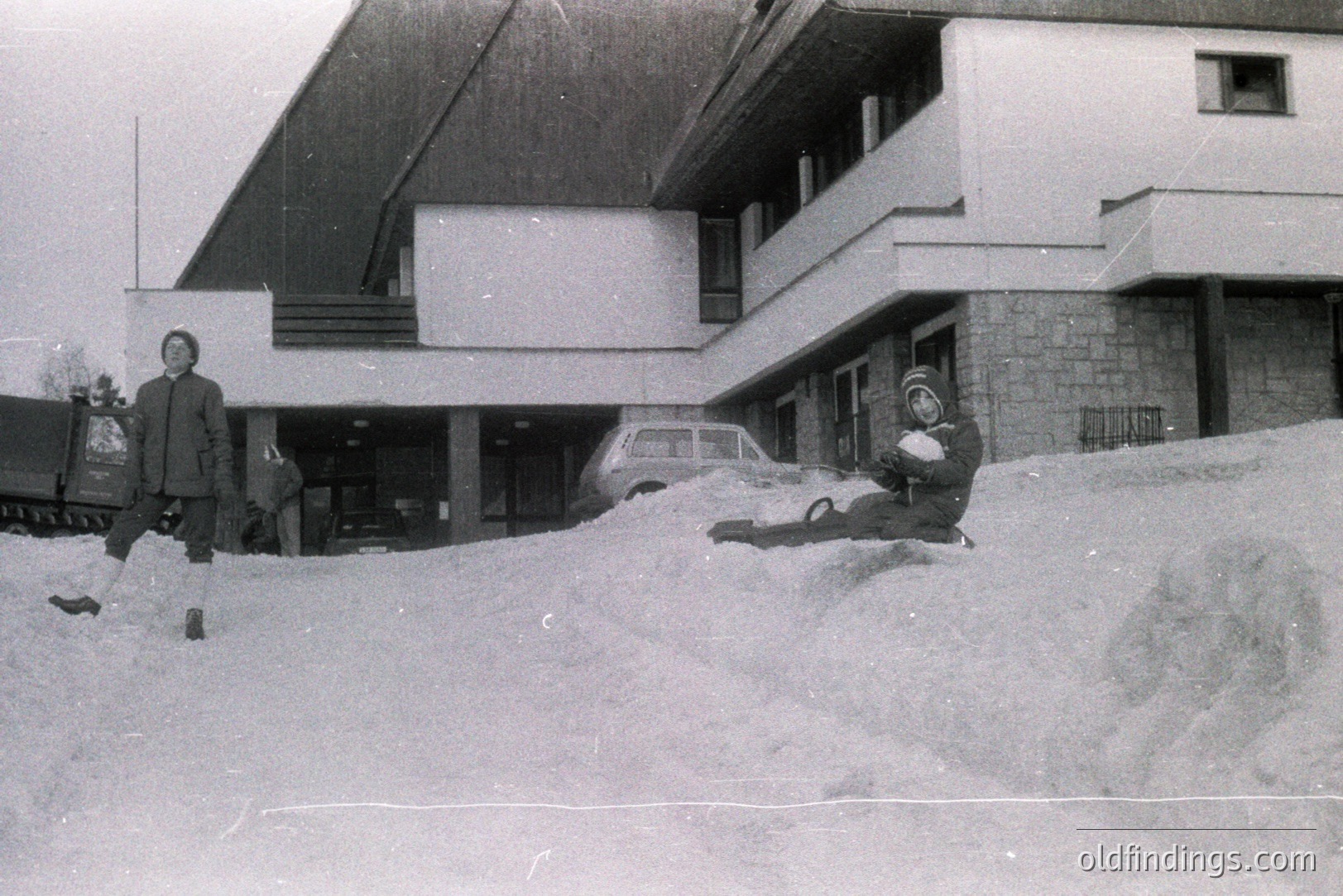 A man in a coat stands in deep snow, observing a child sliding down a large snowbank. The backdrop features a modernist building with prominent cantilevered balconies and stone pillars. Likely a resort or public building. Appears to be Eastern Europe, possibly 1970s.