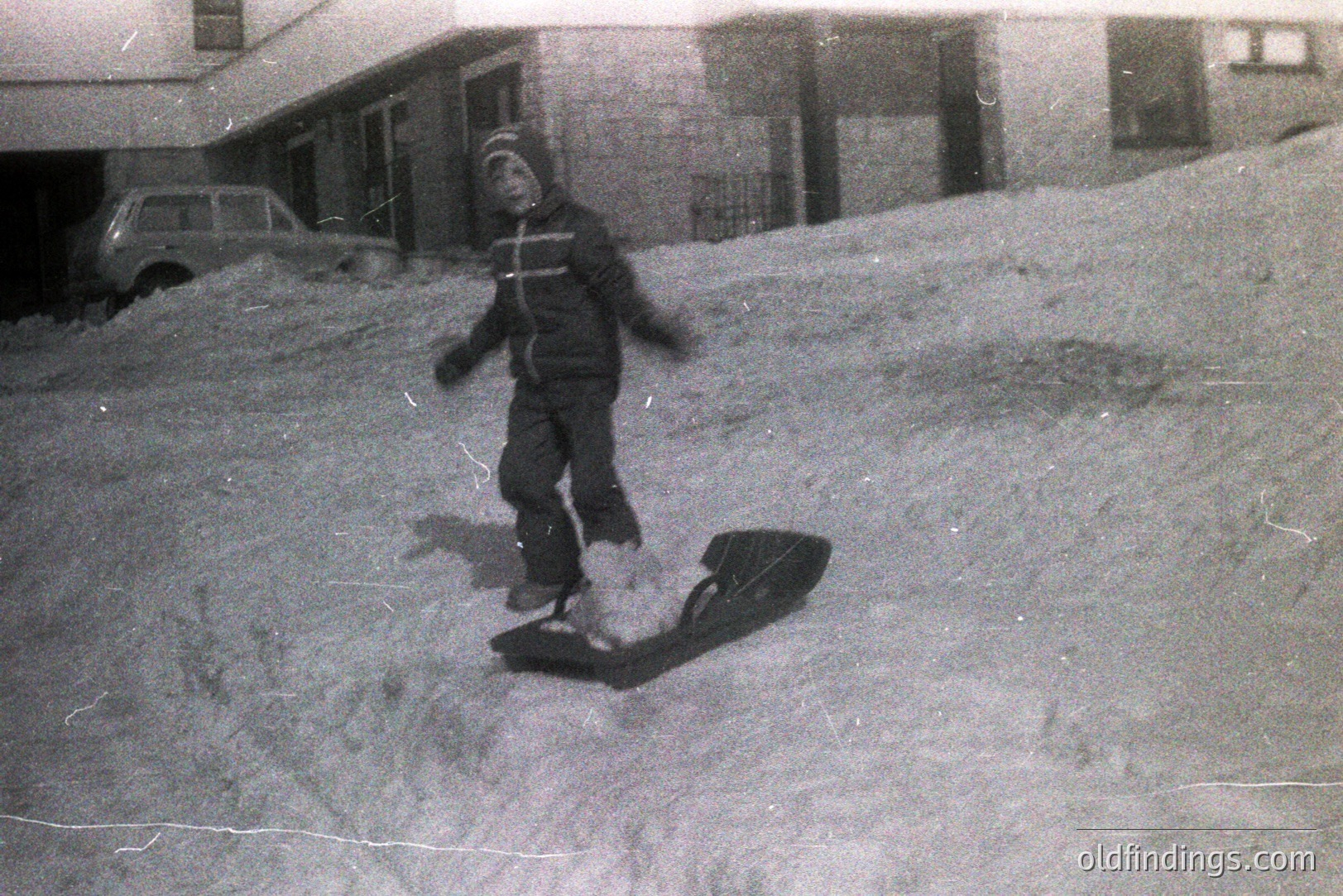 A young child, bundled in a snowsuit and hat, stands next to a wooden toboggan on a snow-covered slope. A vintage car is visible in the background, along with what appears to be a building or structure. Likely mid-20th century, possibly 1950s-1970s. Evokes a nostalgic winter scene.