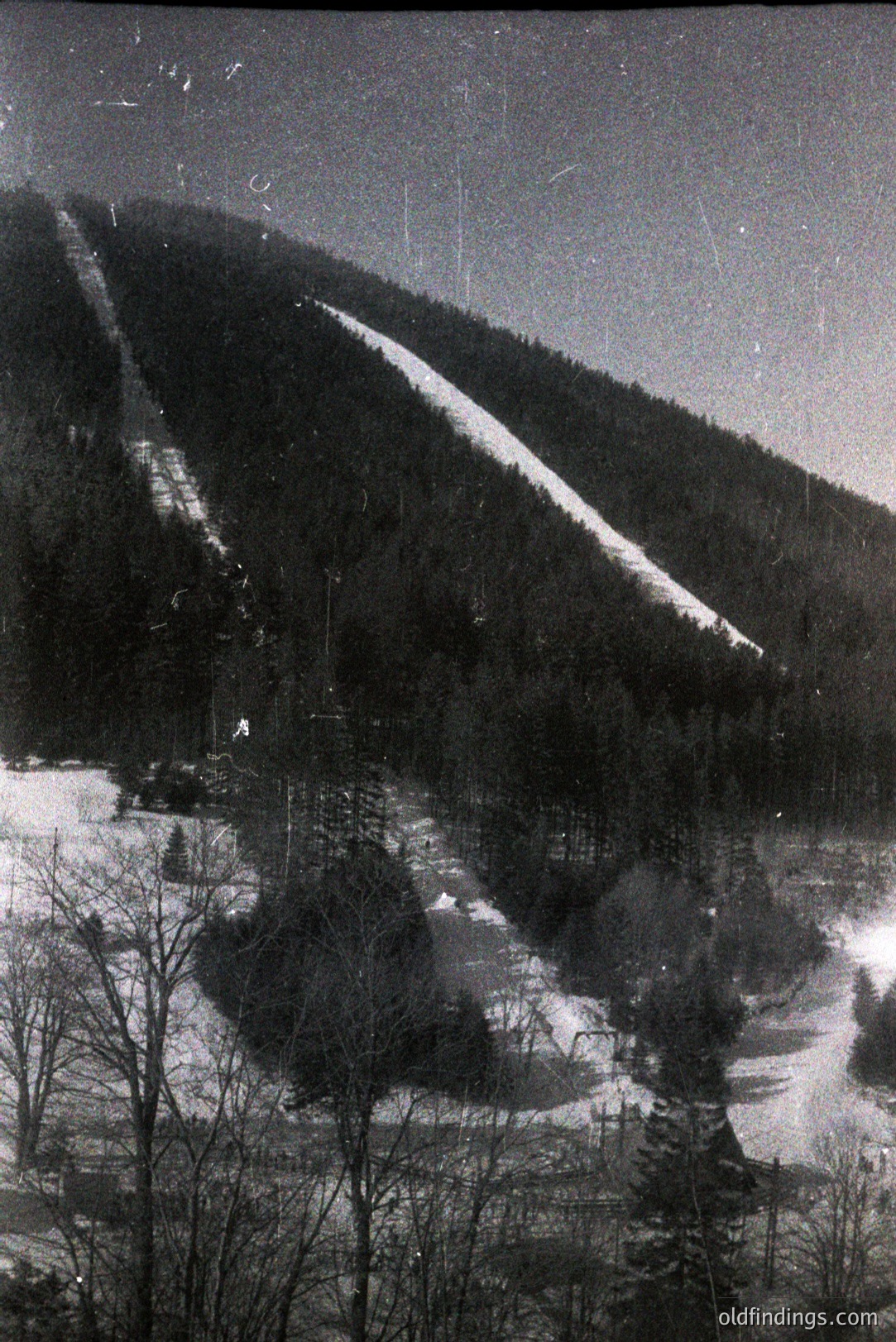 Monochrome landscape captures a snow-covered hillside with a marked ski run. Dense evergreen forest flanks the slope, leading down to a valley filled with snow. Structures visible in the midground hint at a ski resort or rural settlement. Likely 1950s-1970s, possibly Eastern Europe. Potential value for archival, historical, or design referencing.