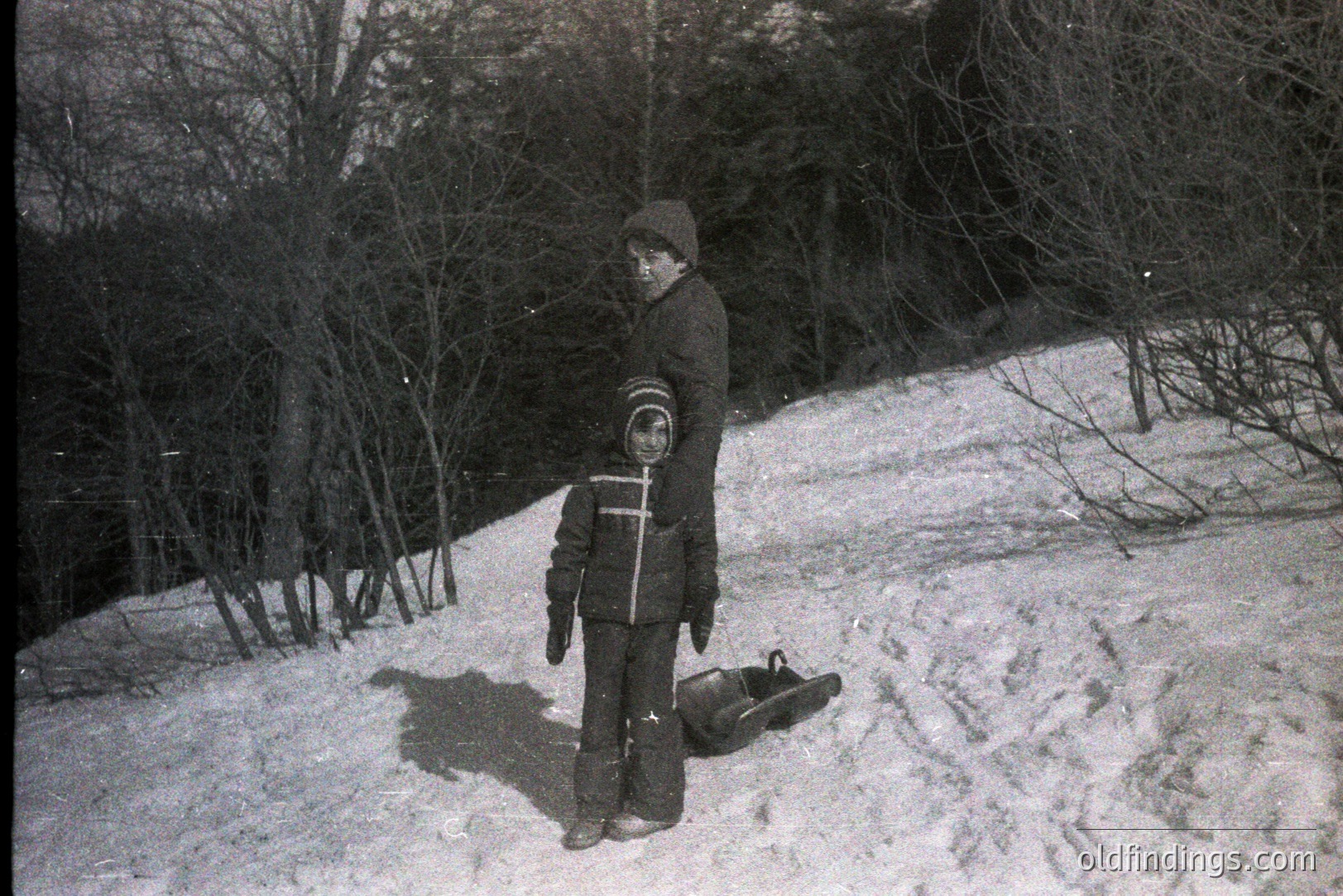 A young boy stands on a snow-covered slope, bundled in a thick winter coat, hat, and snow pants. A wooden toboggan rests at his feet. Sparse trees form the wooded background, typical of a rural winter scene. Likely a candid snapshot, circa 1960s-1970s.